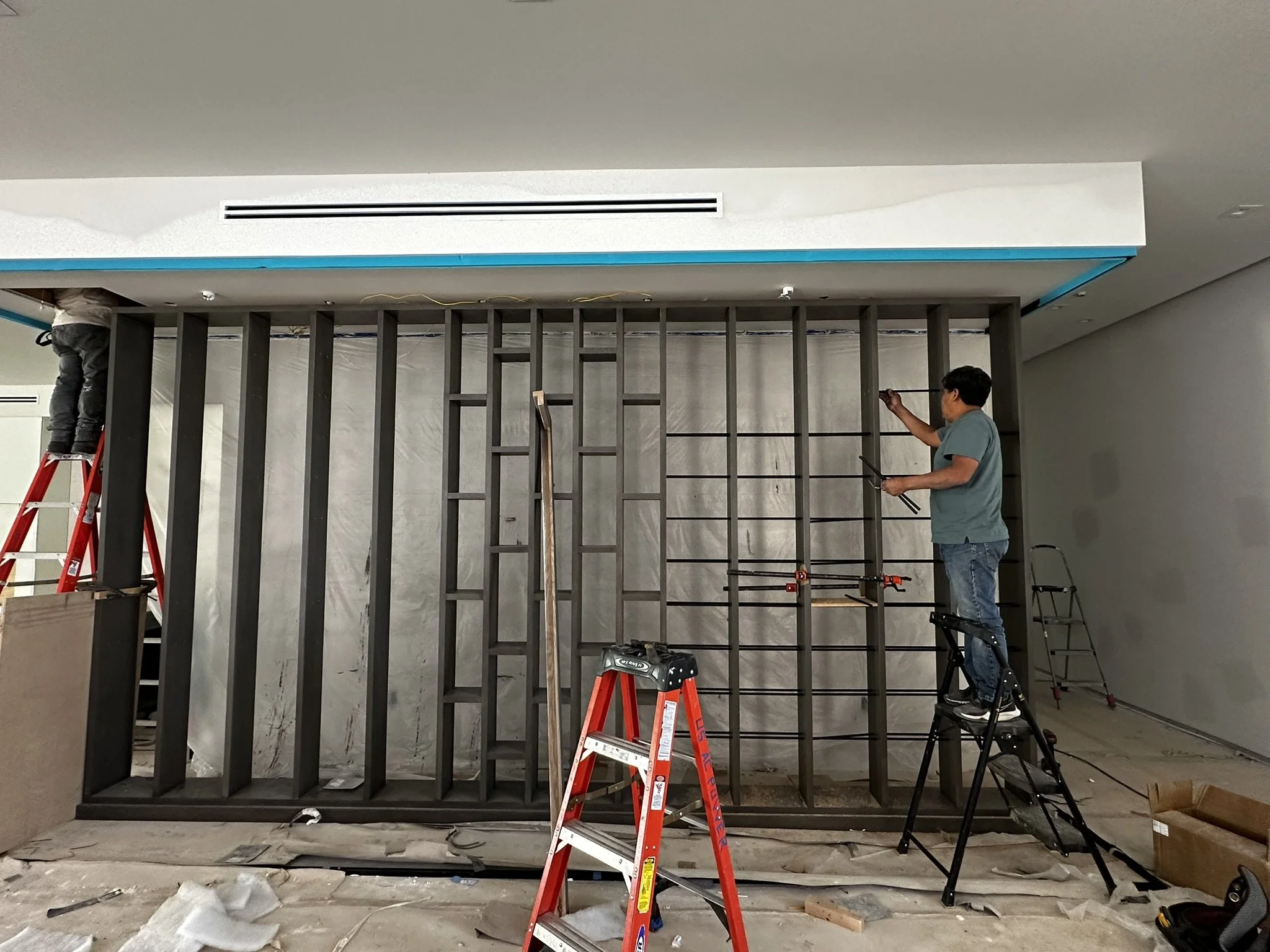 Two workers are installing a large wall or room divider, with the structure's vertical beams visible. One worker is standing on a ladder on the left, while the other is standing on a small platform on the right, both focused on the installation. Cons