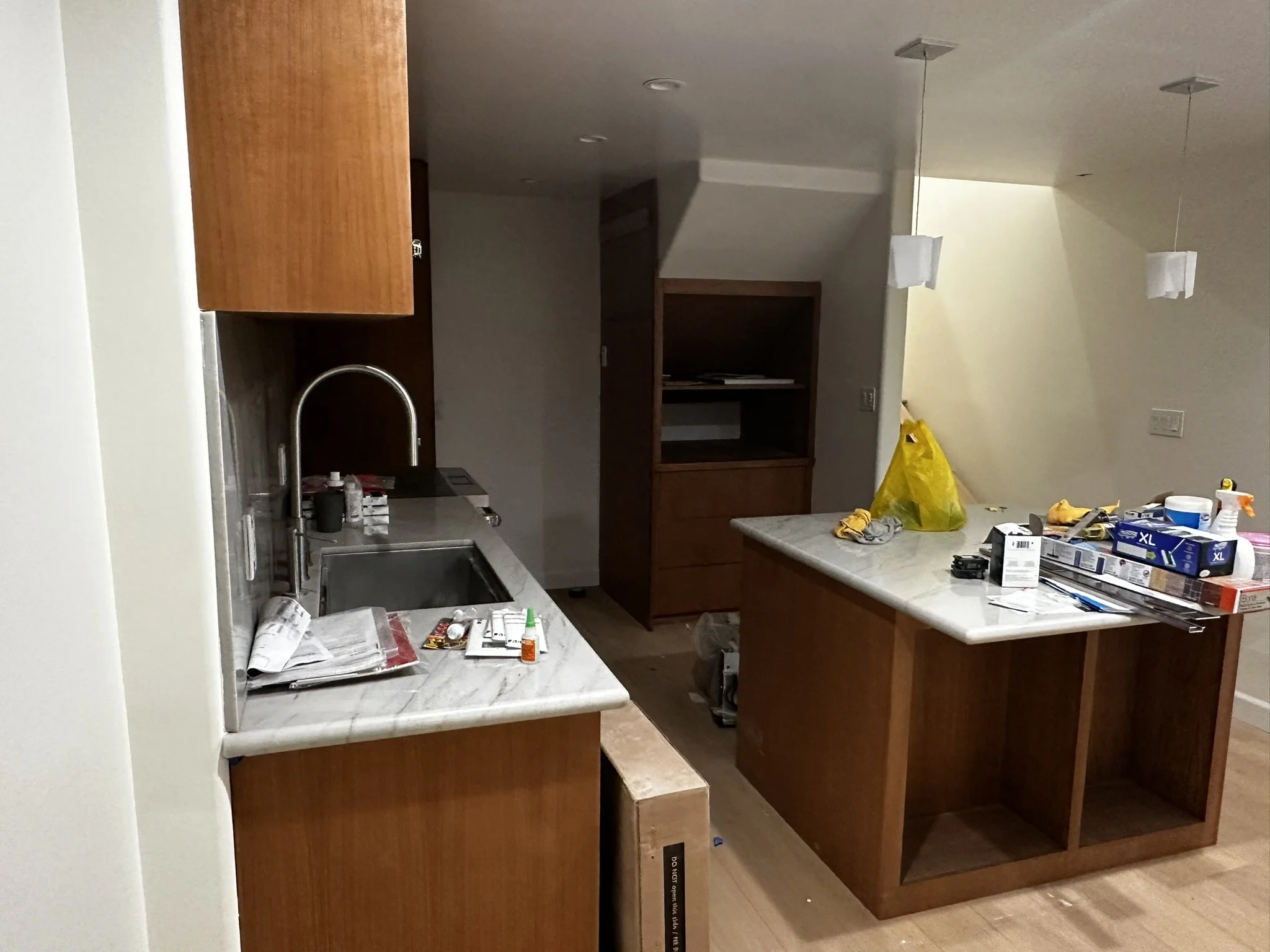 Kitchen with wooden cabinets, marble countertops, and various items including paper, glue, and boxes on the counter, with an open shelf and hanging pendant lights.