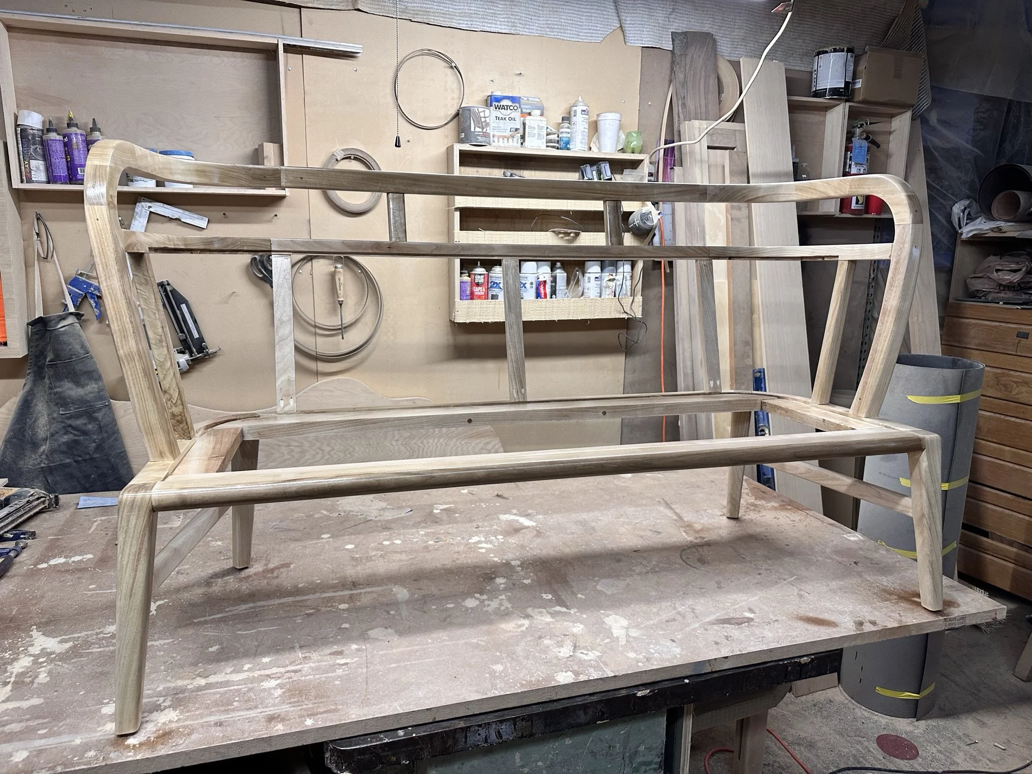 A wooden bed frame under construction in a woodworking workshop with tools and supplies on shelves in the background.