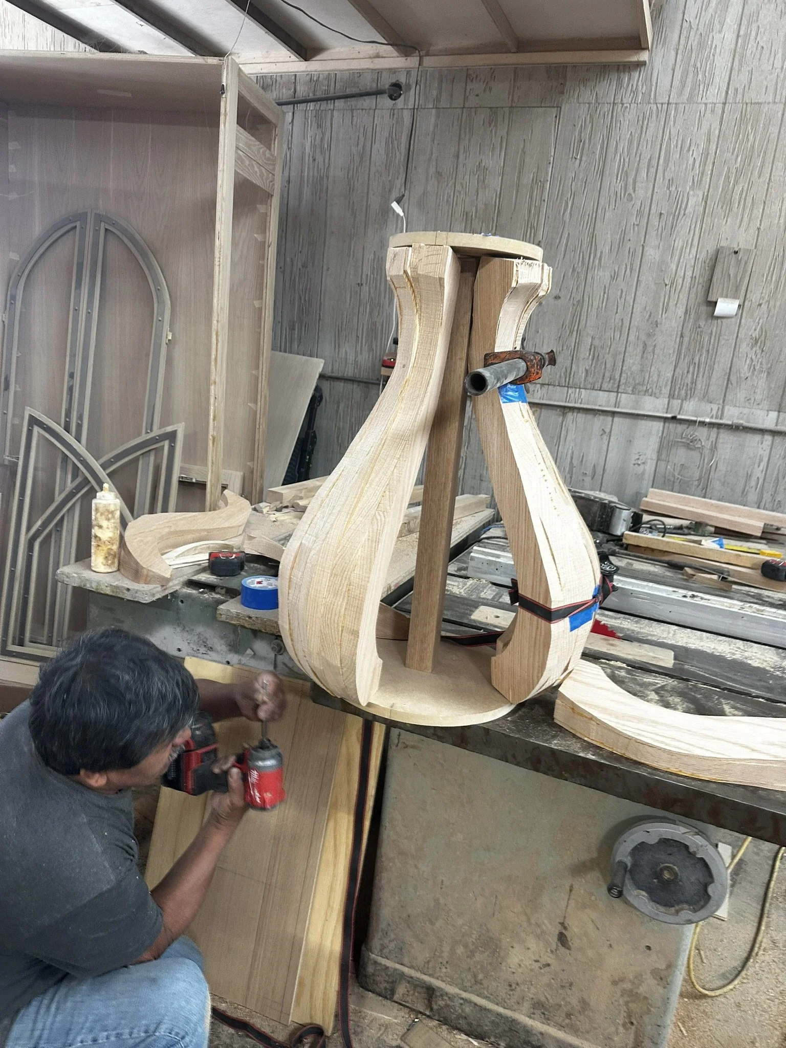 A person working on a wooden chair frame in a woodworking workshop. The person is using a drill and the chair frame is mounted on a workbench.