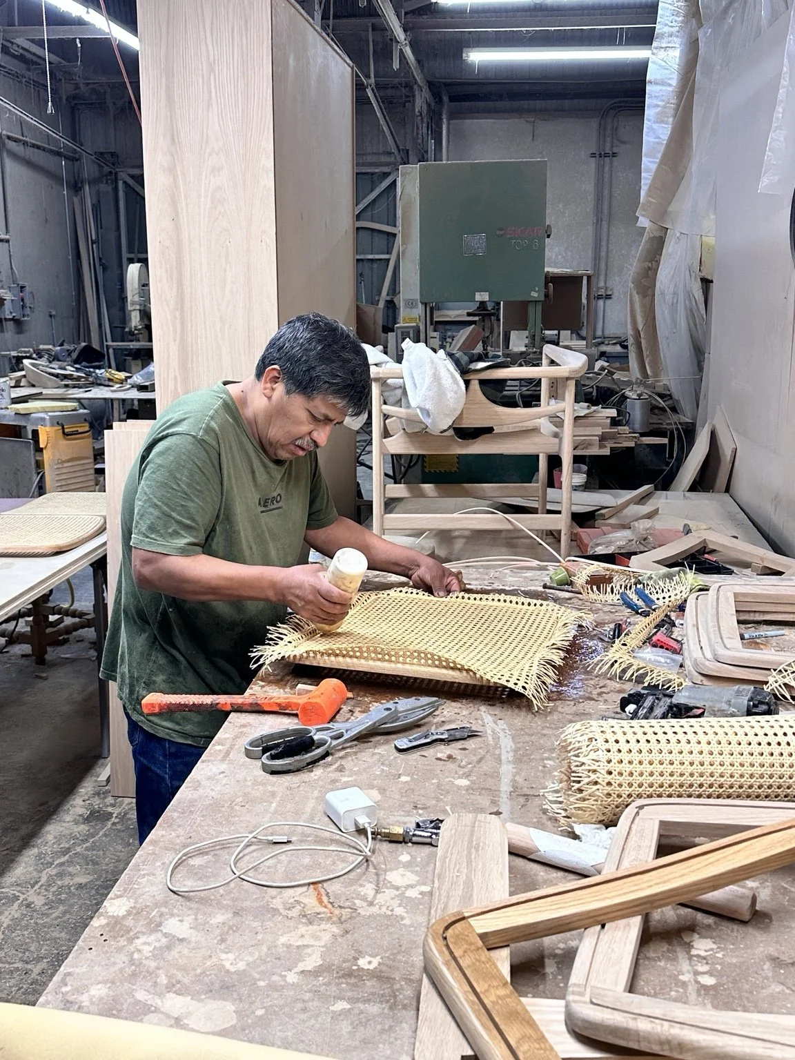 A man working on a craft project at a cluttered woodworking workshop, applying glue to a woven cane seat. The workspace is filled with tools, wooden frames, and equipment.