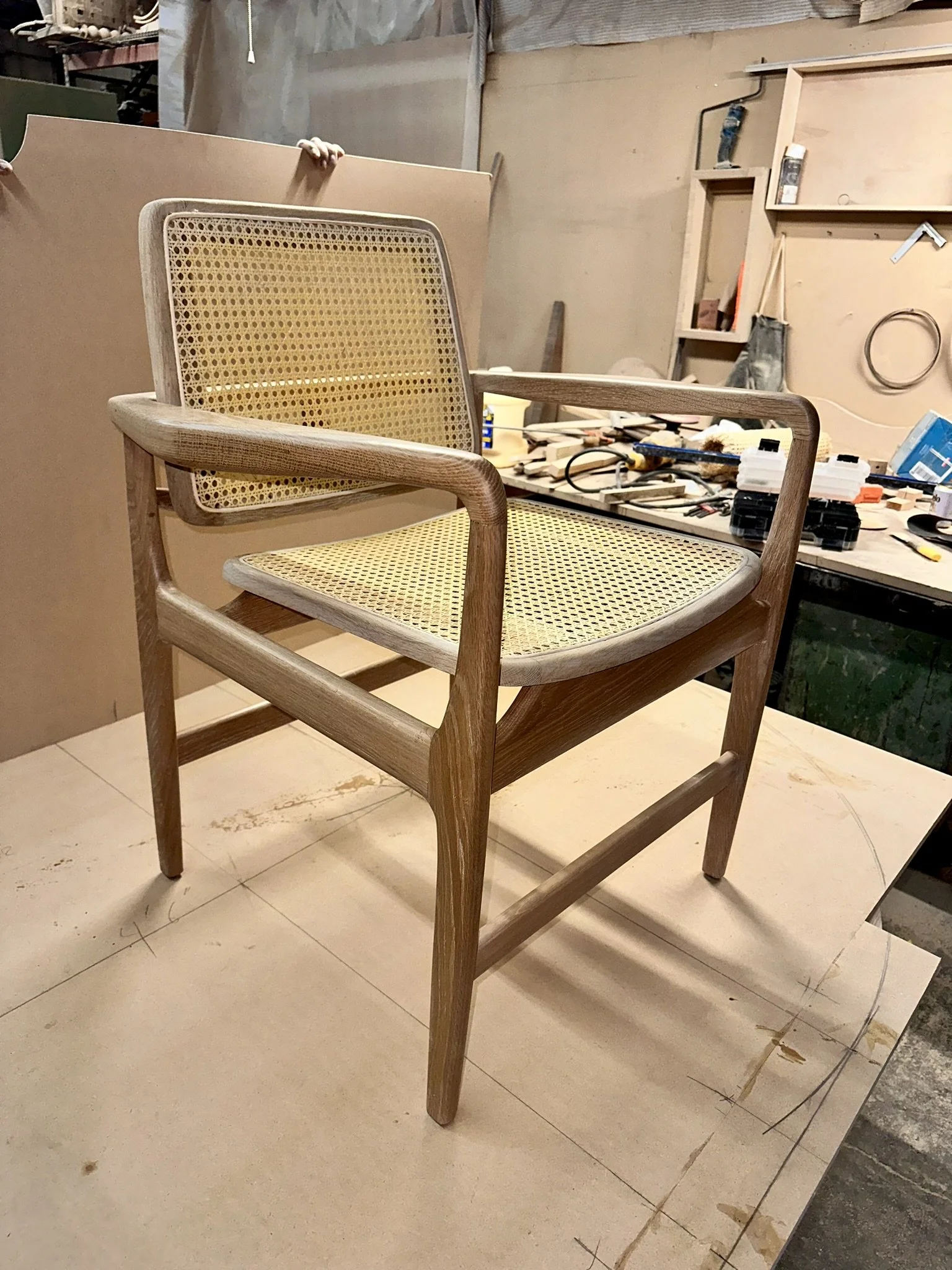 A wooden chair with woven cane backrest and seat positioned on a beige tiled floor in a workshop or carpentry space with tools and wooden materials visible in the background.