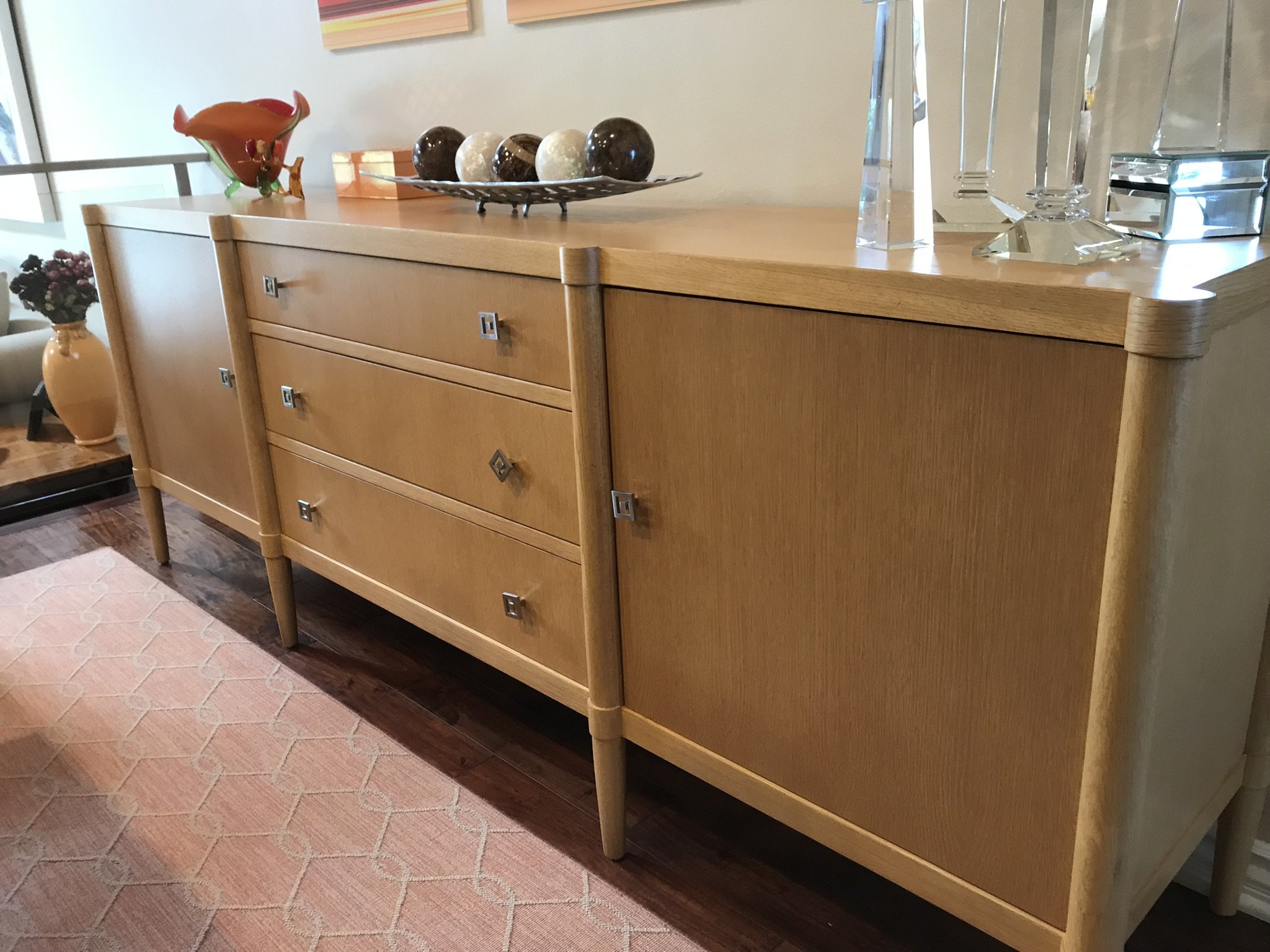 A mid-century modern wooden sideboard with four drawers, decorative square handles, and slightly rounded edges. It sits on four tapered legs. On top, there are decorative items including a glass sculpture, a bowl of spherical objects, and a piece of 