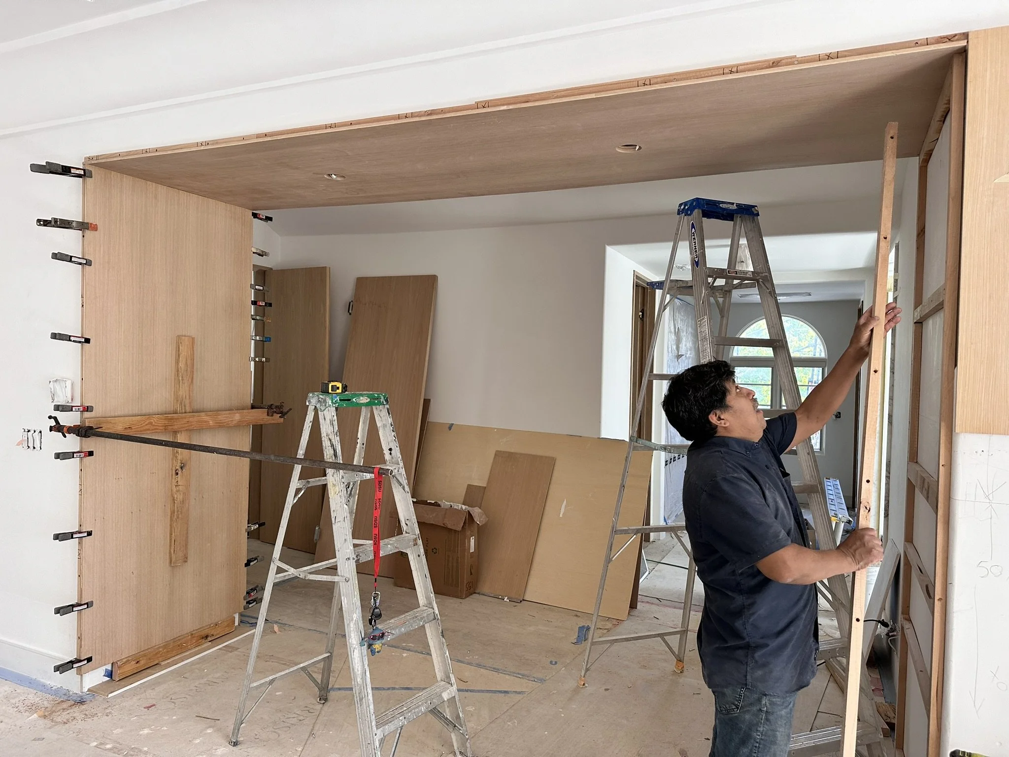 A person working on installing a wooden partition or wall indoors, using a stepladder, clamps, and tools, with some wood panels and construction materials visible around.