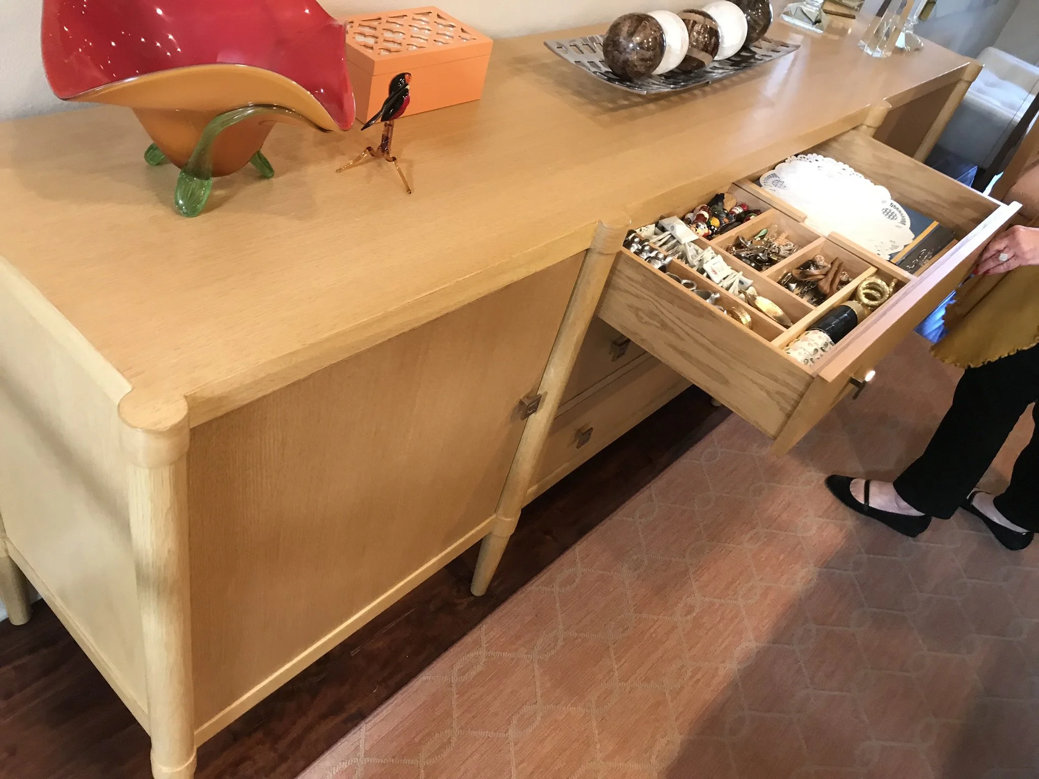 A wooden dresser with a drawer partially open, revealing various jewelry items, and decorative objects on top, including a red glass vessel with green legs, a glass bird sculpture, and a tray with spherical objects and vases.