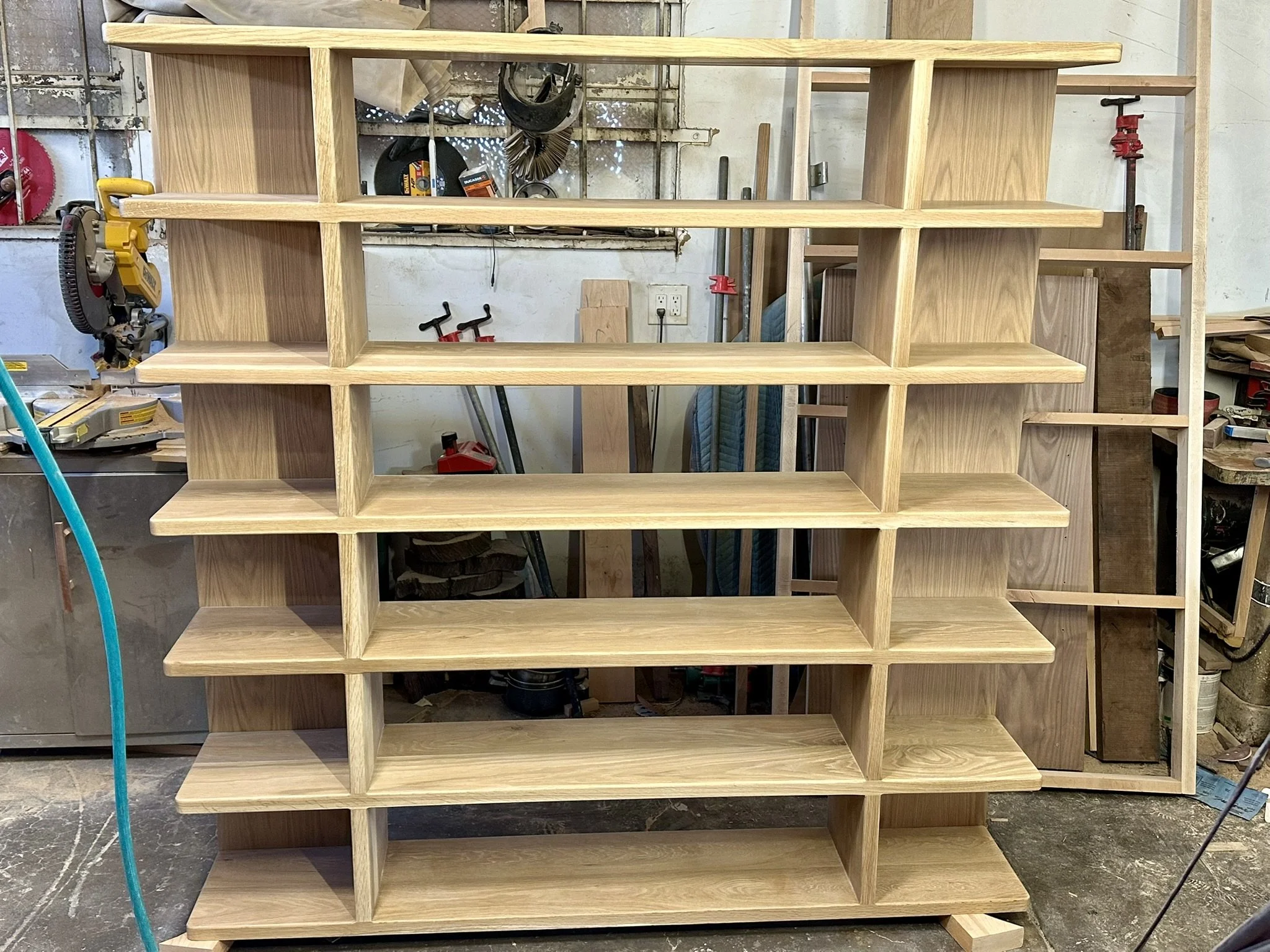 Unfinished wooden bookshelf en madera en un taller de carpintería con varias herramientas y materiales alrededor.