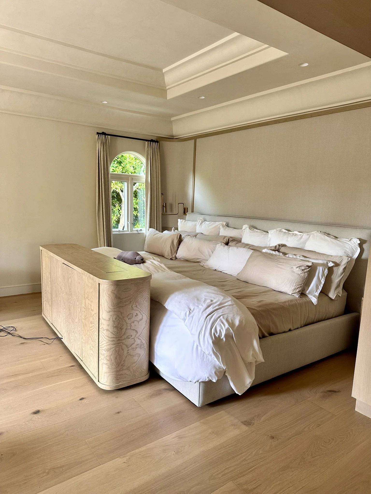 A bedroom with a large bed, white and beige bedding, a wooden nightstand, a window with beige curtains, and light-colored hardwood flooring.