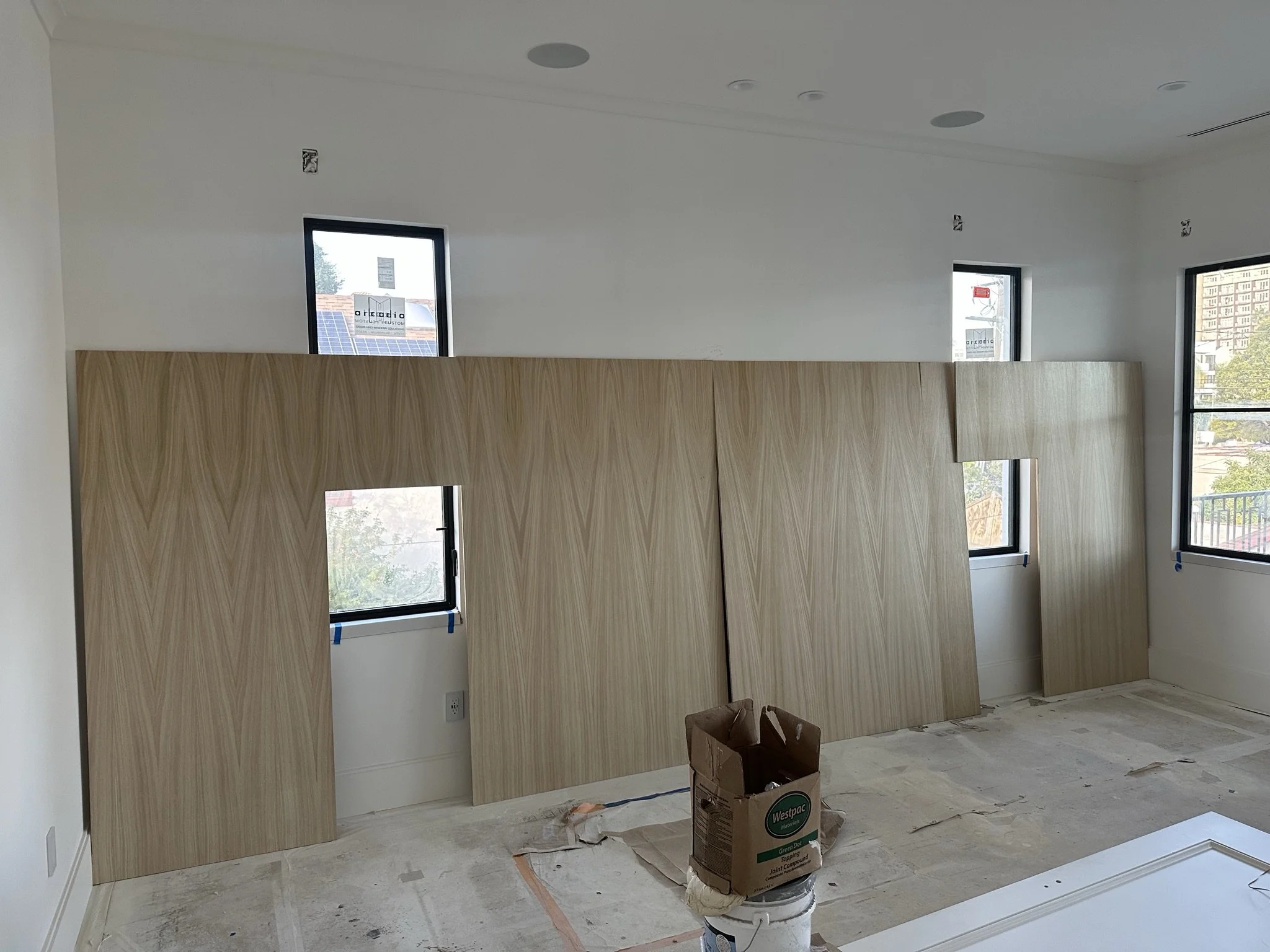 Interior of a room under construction with four wooden panels leaning against the wall, visible windows, and painting supplies on the floor.