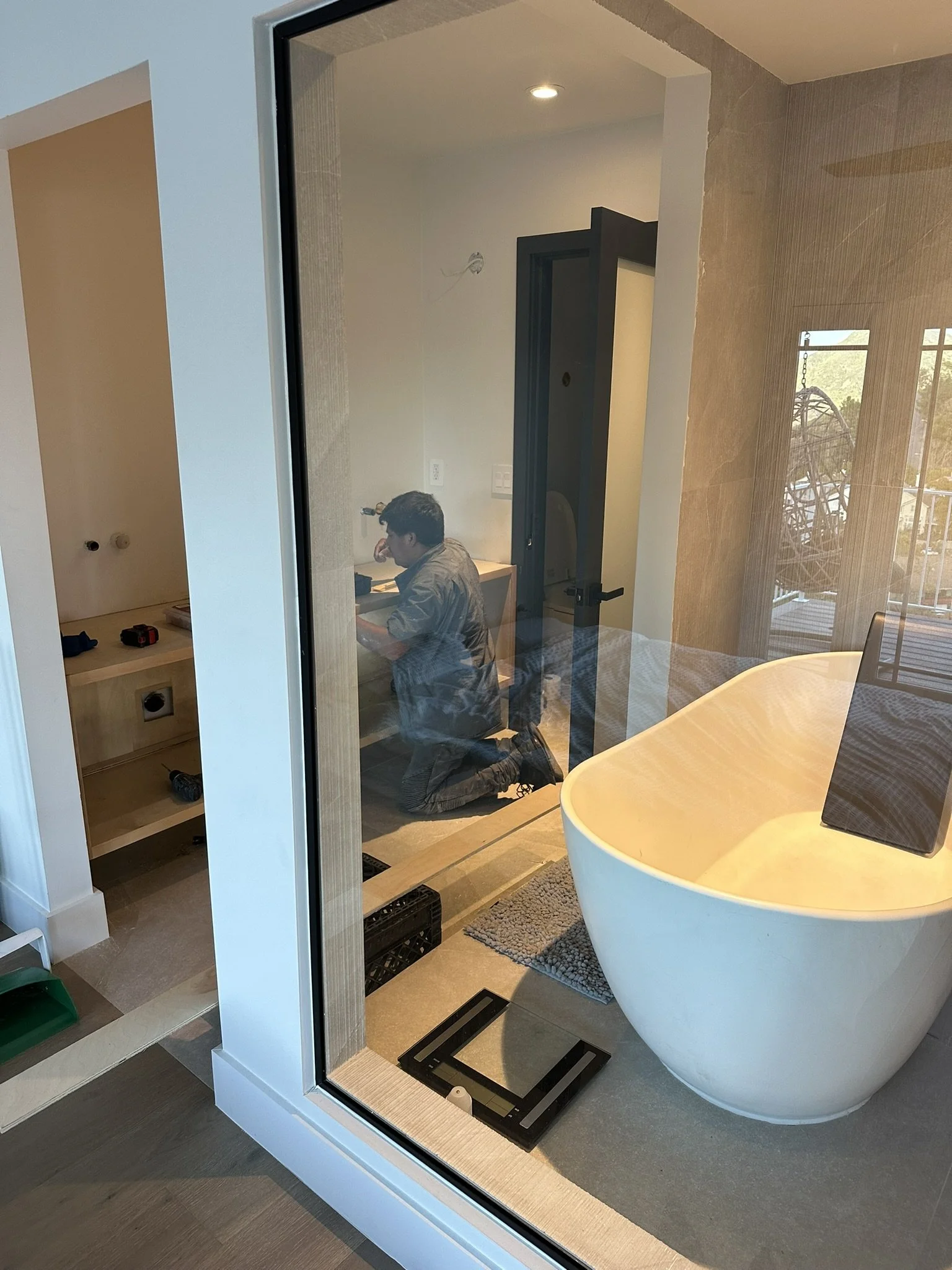 A person kneeling in a bathroom, working on installation or repair, with a large free-standing bathtub, a black scale on the floor, and a glass shower enclosure. Outside the enclosure, a window with trees outside.