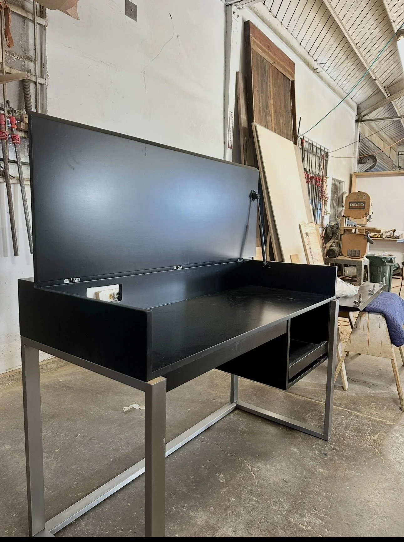 A black and gray metal fire pit or outdoor stove with a hinged lid in a garage or workshop, with woodworking tools and materials in the background.