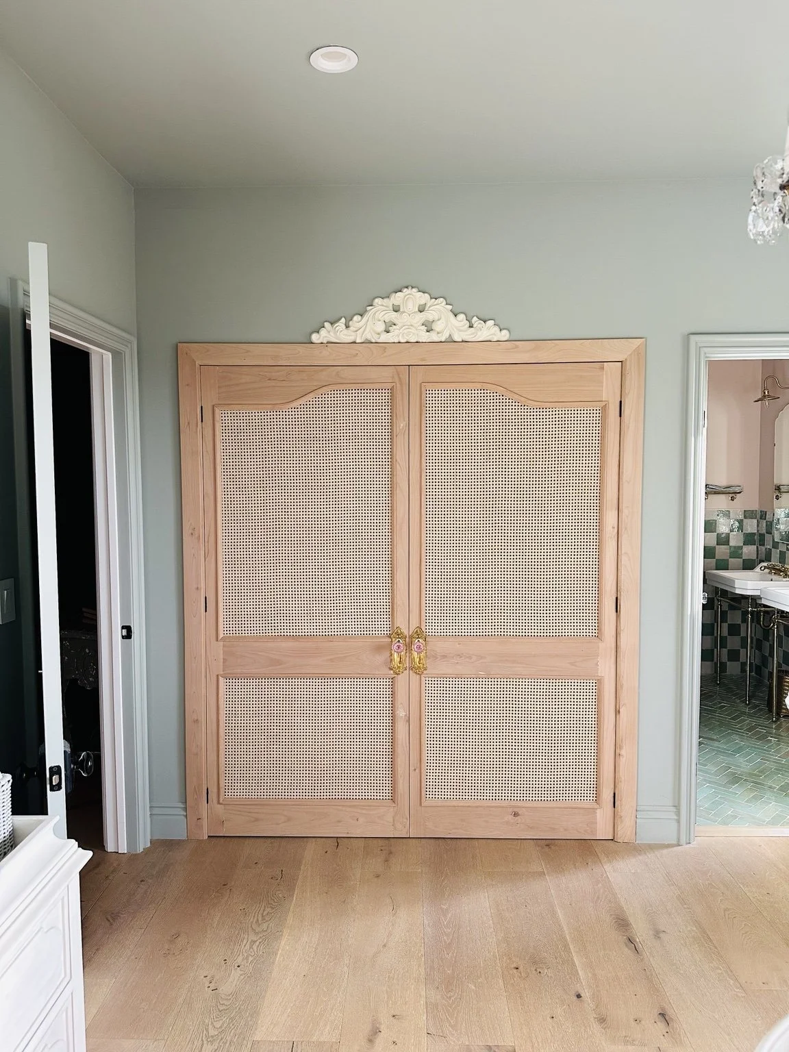 A wooden cabinet with cane doors and ornate gold handles, placed against a light green wall. There is decorative white molding above the cabinet. To the right, there is a bathroom with green and white tiles and a sink. To the left, part of a doorway 