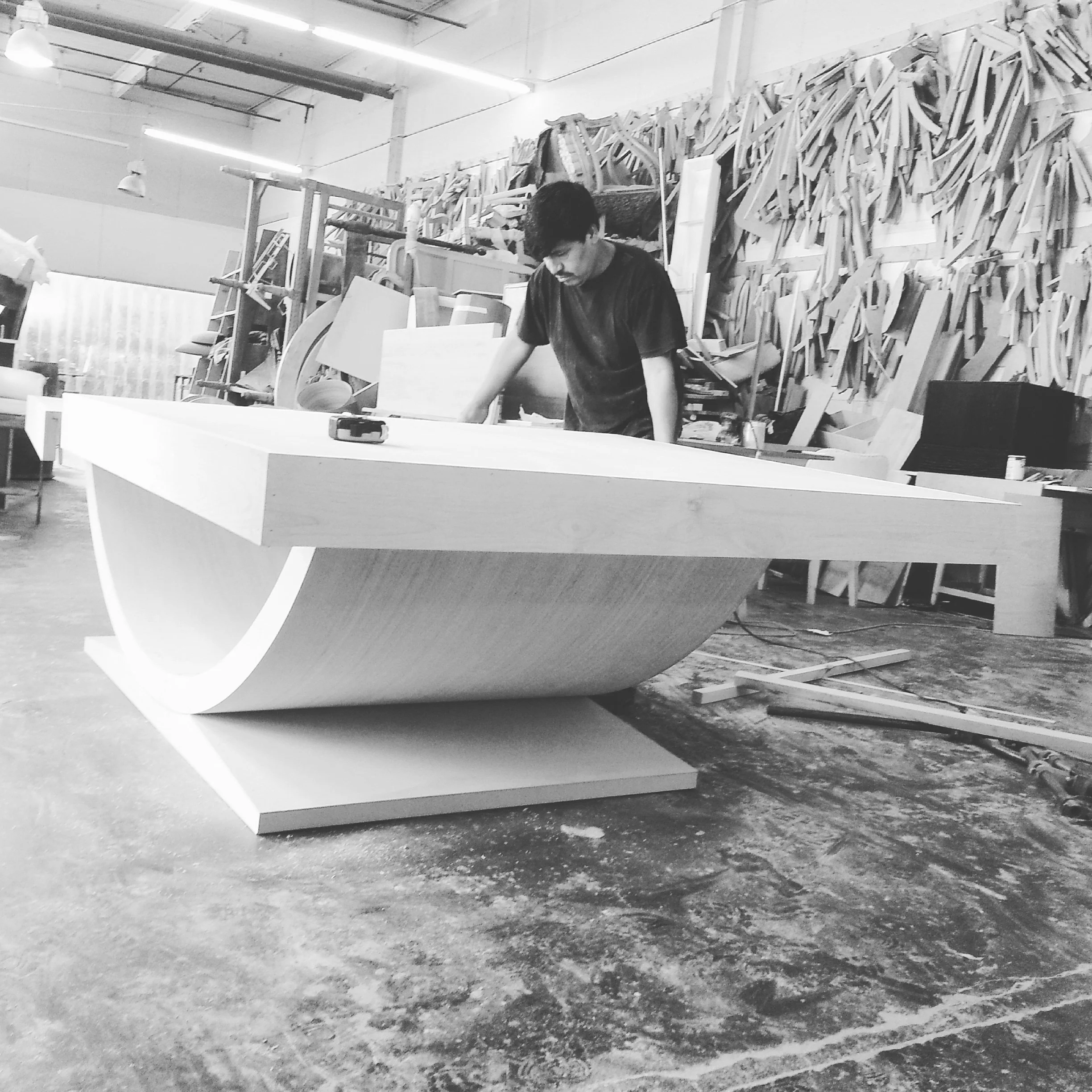 A person working on a large curved white object in a woodworking workshop, with wooden materials stacked on the wall behind.