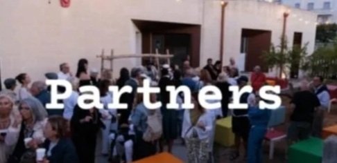 Gruppo di persone che socializzano e partecipano a un evento all'aperto, con tavoli colorati e una palizzata sullo sfondo, scritto 'Partners' in sovrimpressione