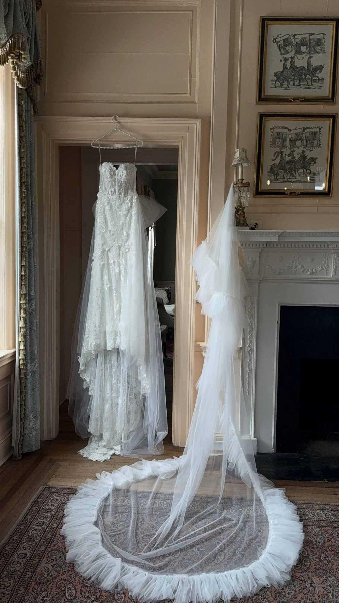 A wedding dress hangs on a hanger in a room with cream-colored walls, next to a fireplace with an ornate mantel. The dress is long, white, and lace, with a veil attached to the hanger that drapes onto the floor. A circular wedding veil with ruffled edges is laid out on a rug in front of the dress.