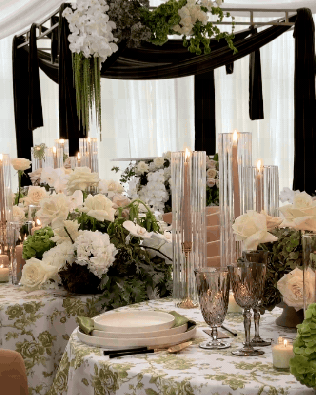 Elegant table setting with white and pale pink flowers, tall glass candle holders, and candles in a decorated indoor event space.