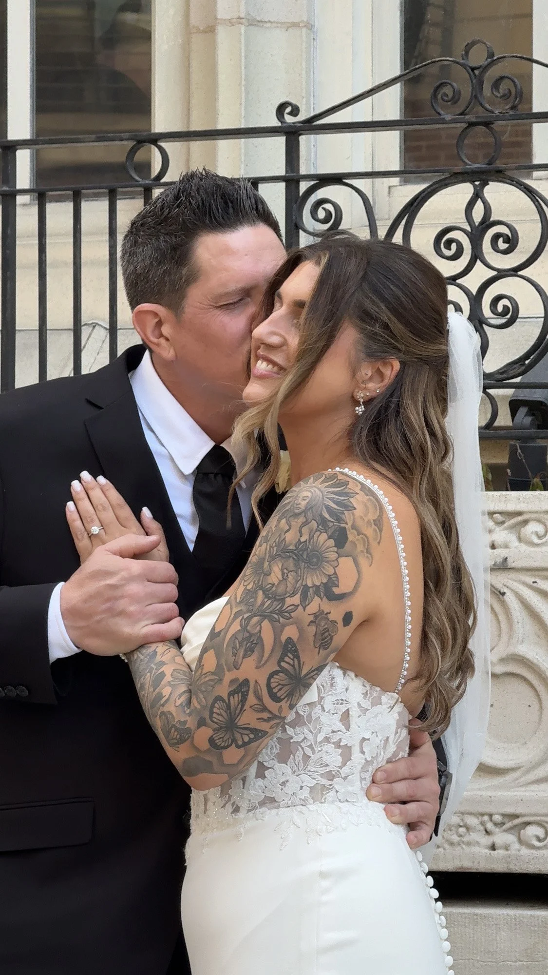 A couple dressed in wedding attire sharing a kiss outside a building, with a metal fence in the background. The woman has long wavy hair, tattoos on her arms, and is wearing a white lace wedding dress. The man has short dark hair, is dressed in a black tuxedo with a white shirt and black tie, and is holding the woman's hand and waist.