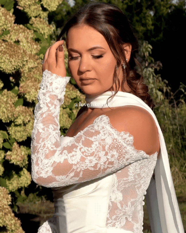 A woman dressed in a white lace wedding gown with an off-the-shoulder design, standing outdoors with greenery in the background, looking down with her hand near her face.