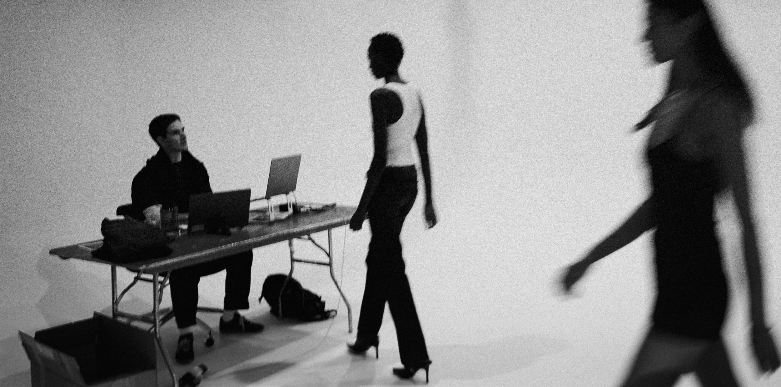 Shawn Dezan, Casting Director, People walking past a photo studio set with a desk, chairs, and laptops in a black-and-white setting.