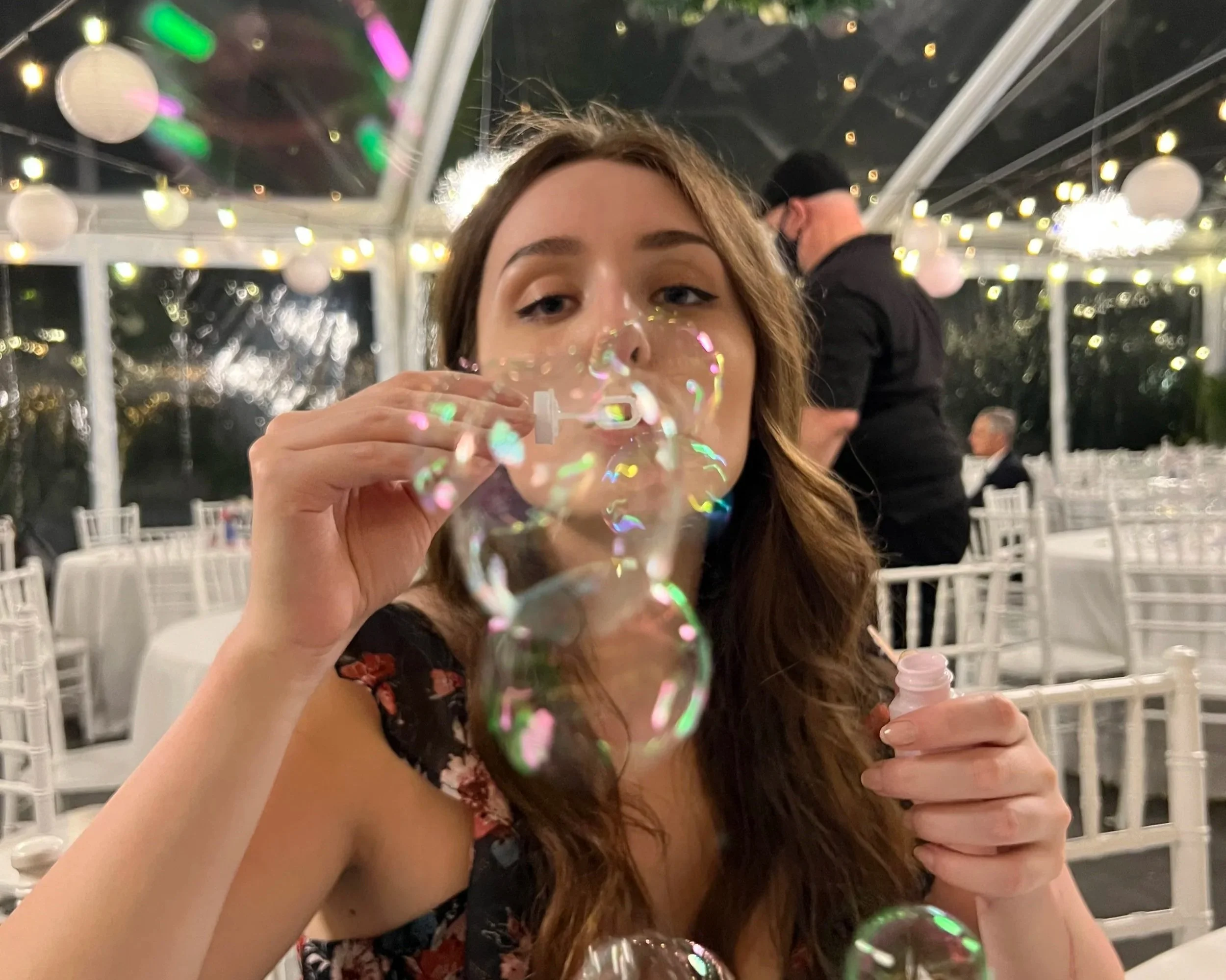 A young woman with long brown hair and wearing a black floral dress blowing bubbles at a formal event, with string lights and white chairs in the background.
