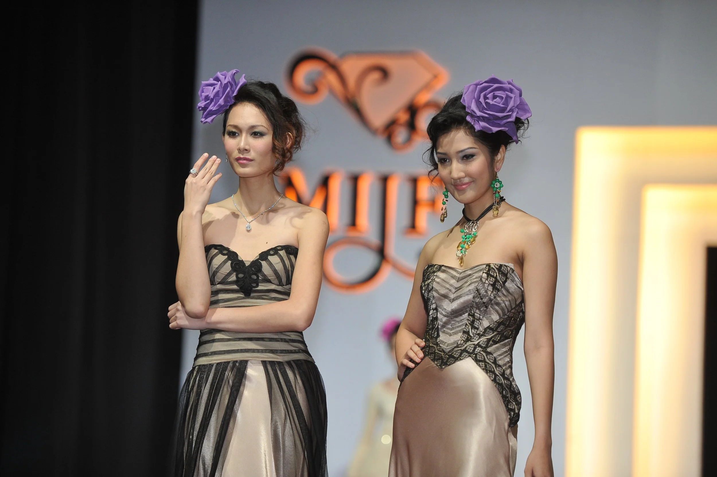 Two women with purple roses on their heads wearing strapless dresses with floral and geometric patterns, standing on a runway with a blurred background.