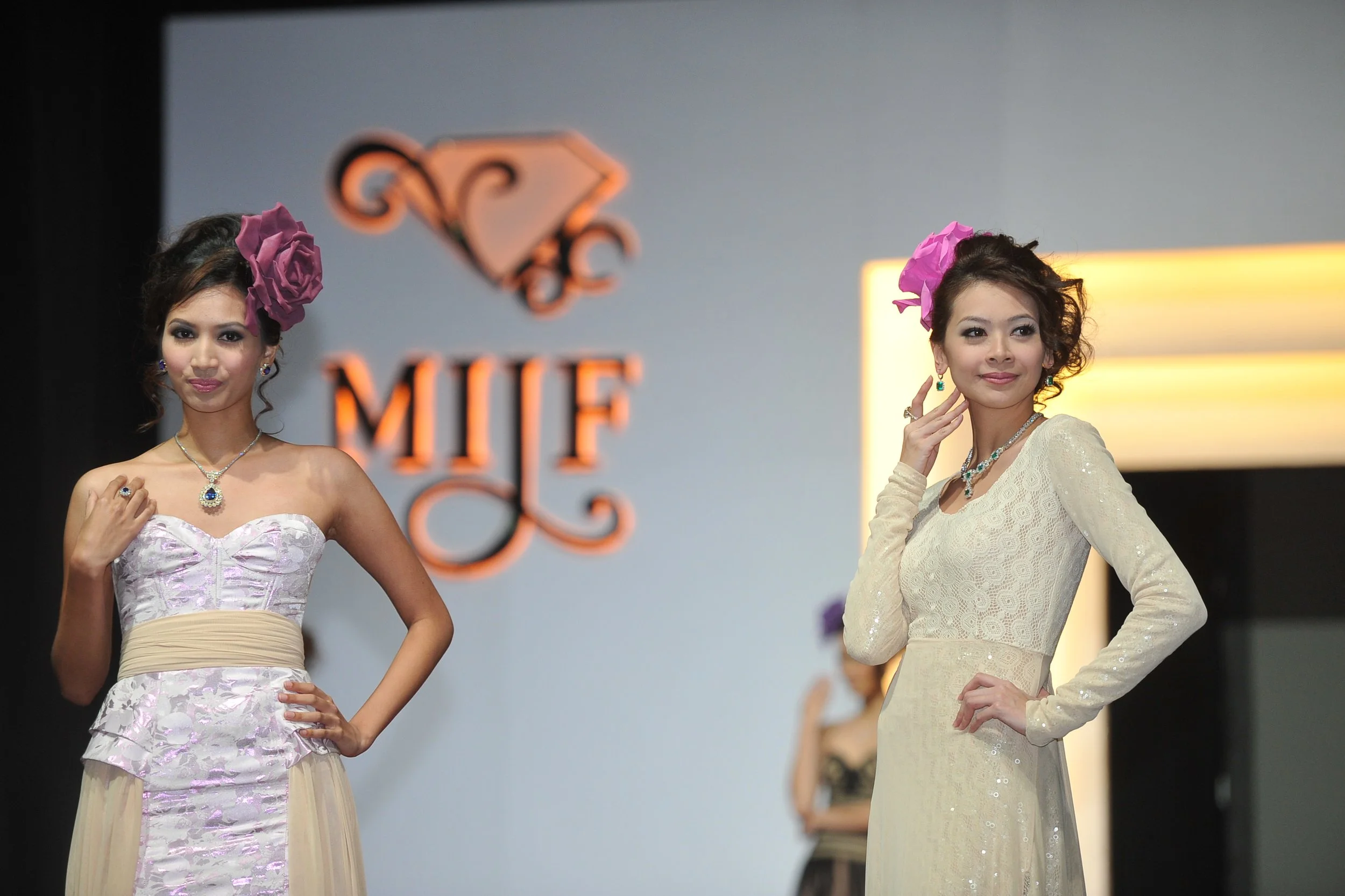 Two women modeling Supreme Gems jewelry on a runway, wearing jewelry and floral headpieces, with a blurred designer logo in the background.