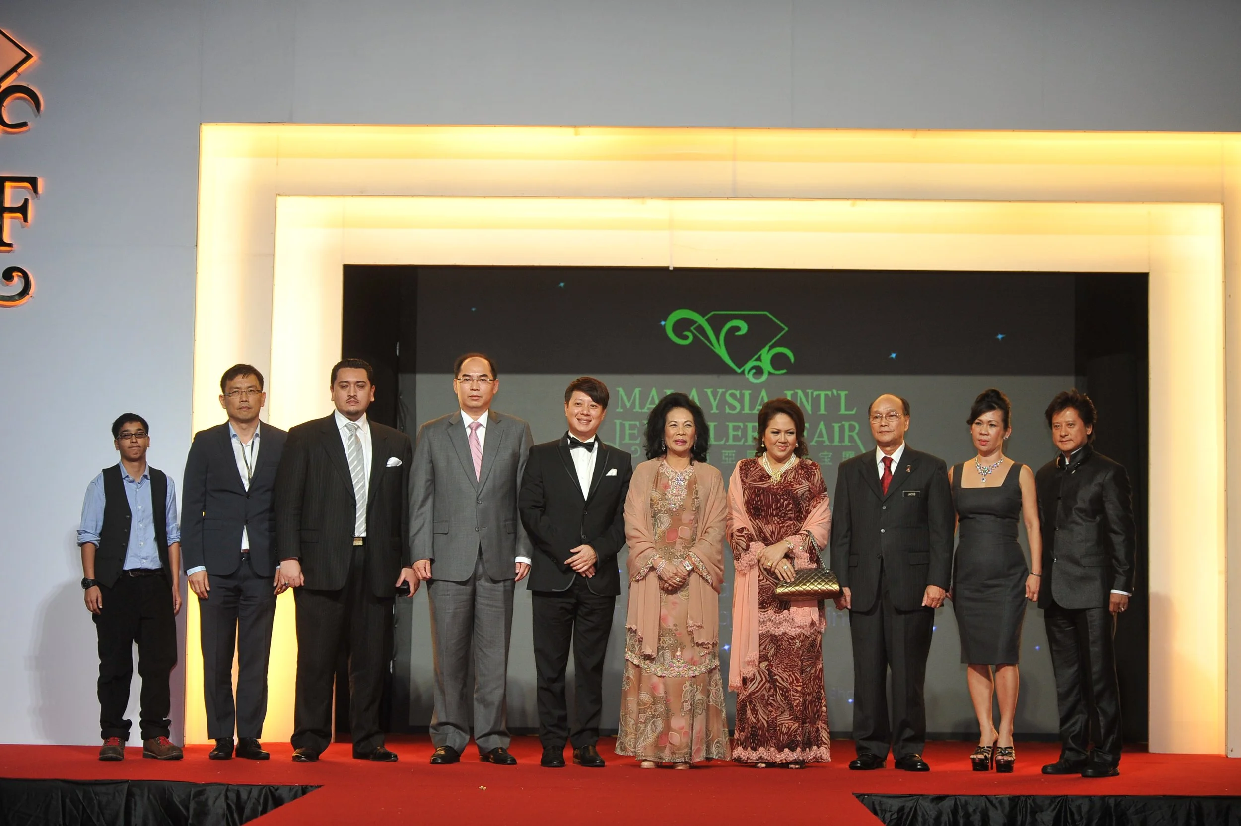 A group of men and women dressed in formal attire standing on a stage with a red carpet in front of a backdrop that reads "Malaysia International Jewelry Fair" in green, with a logo of a stylized leaf or jewelry design above it.