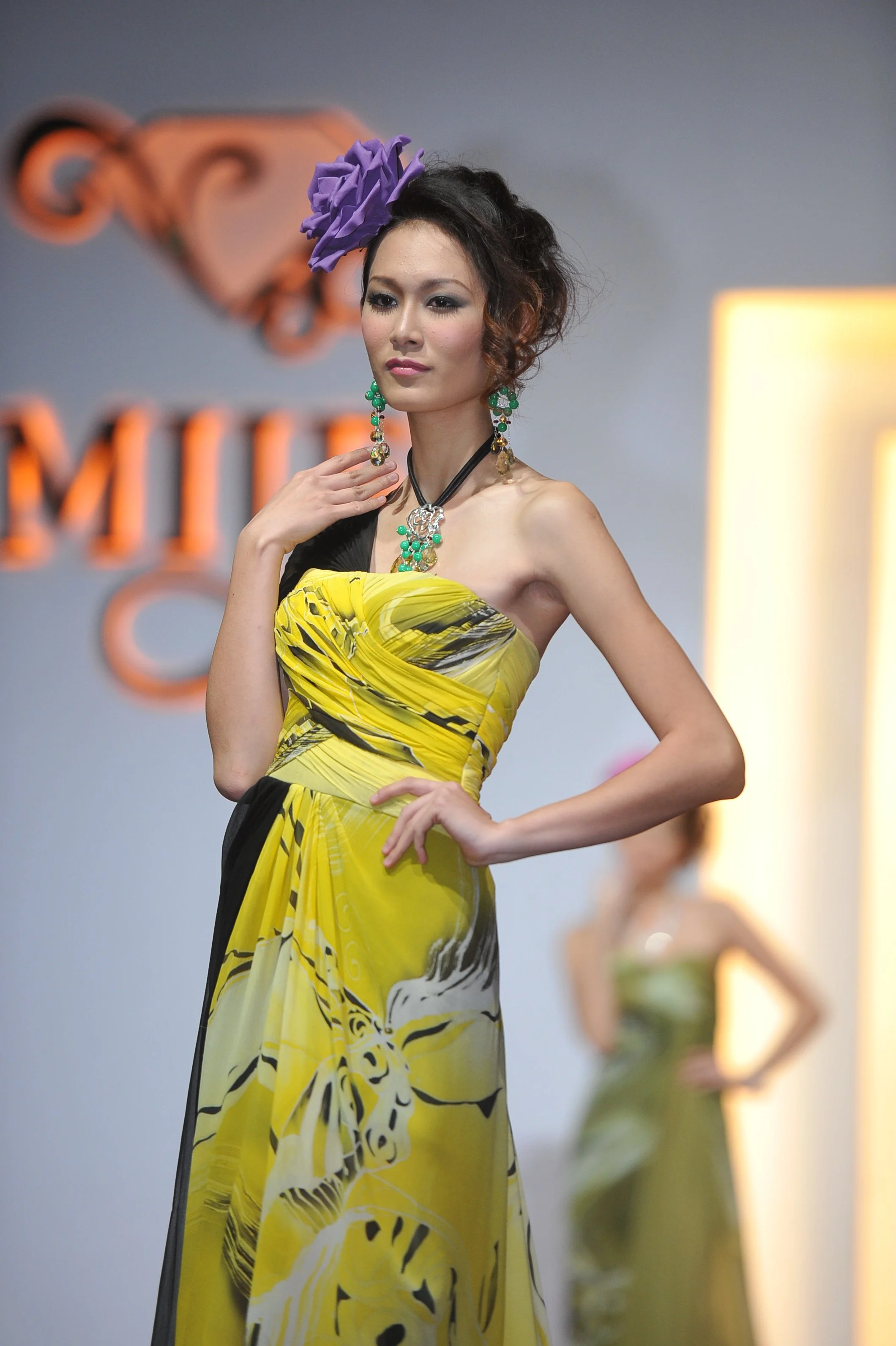 Fashion model on runway wearing yellow and black strapless dress with purple flower hair accessory and statement earrings.