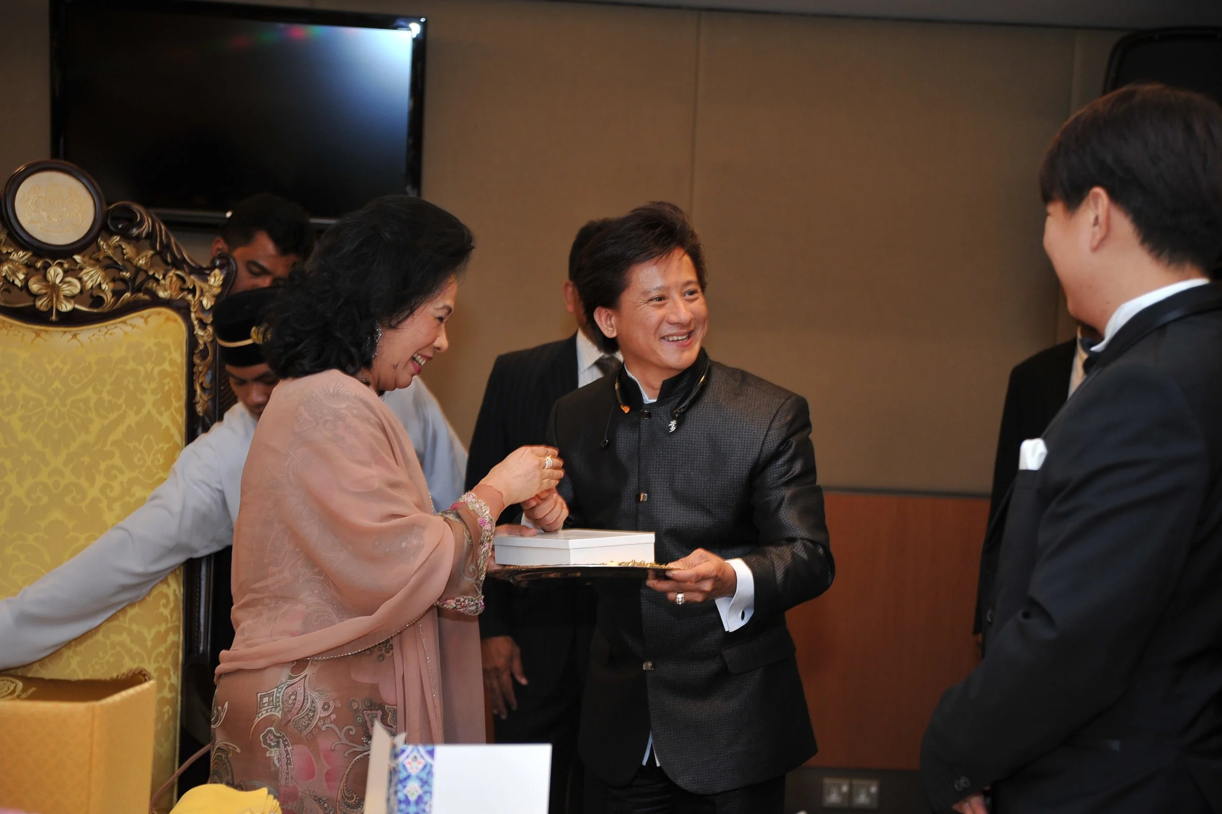 A group of people, including a woman in traditional attire, are smiling and exchanging what appears to be a gift or an award in an indoor setting with a decorated yellow chair in the background.
