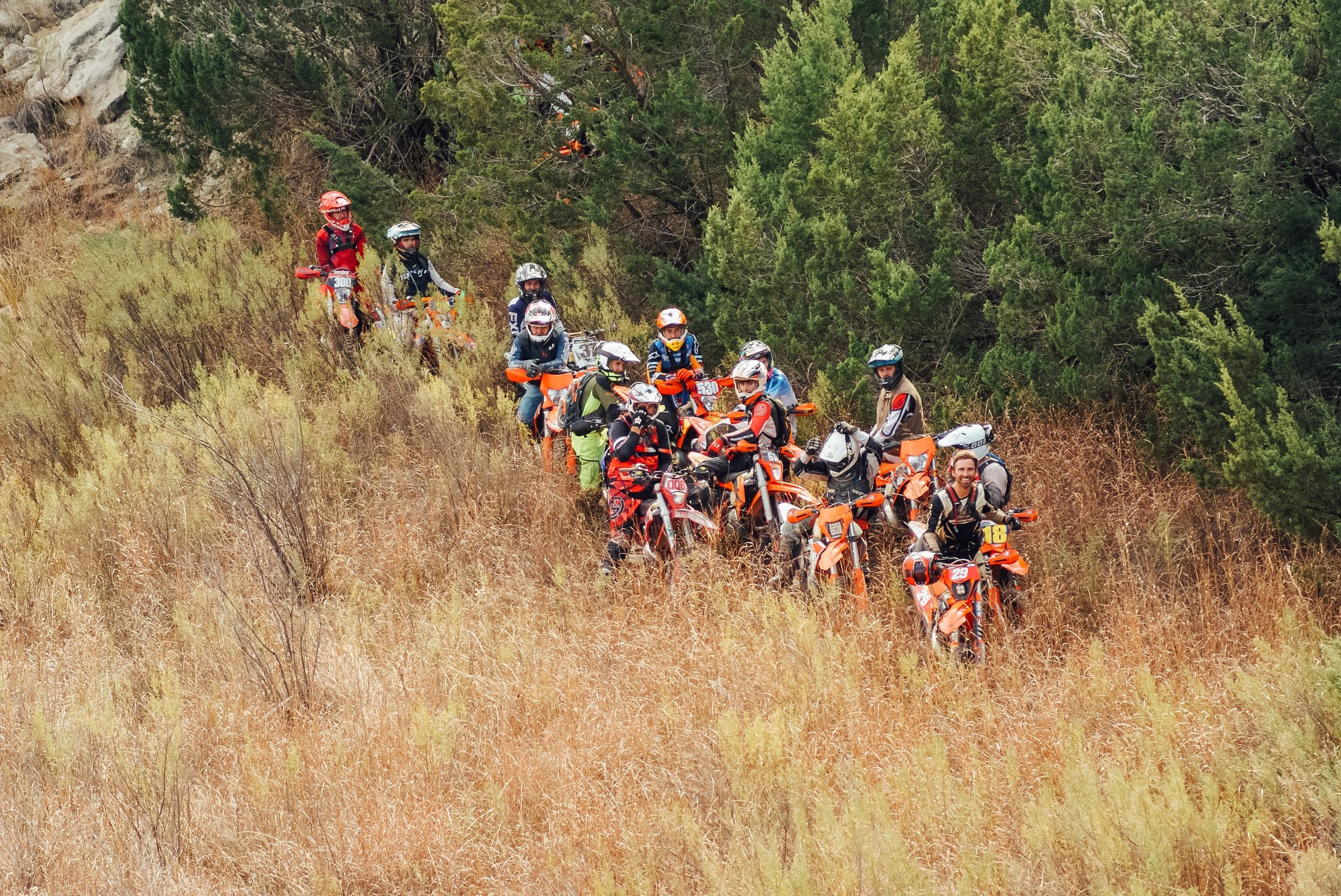 Group of motorcyclists, wearing helmets and gear, riding on a dirt trail through a grassy and wooded area.