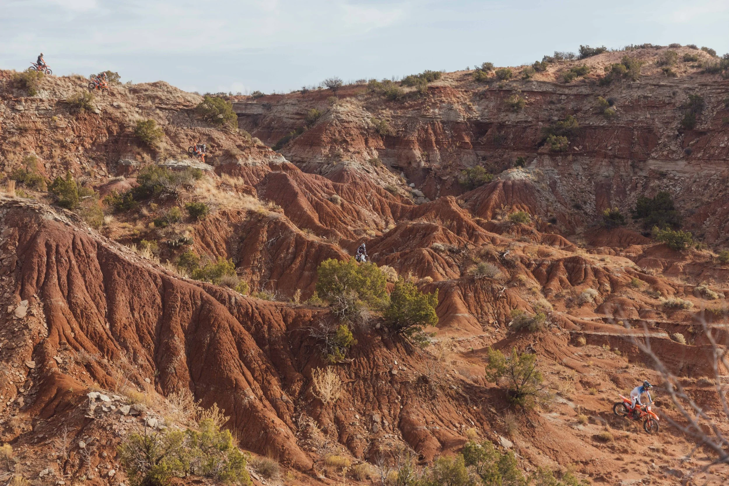 Off-road motorcycle riders on a rugged, reddish-brown canyon landscape with sparse vegetation under a cloudy sky.