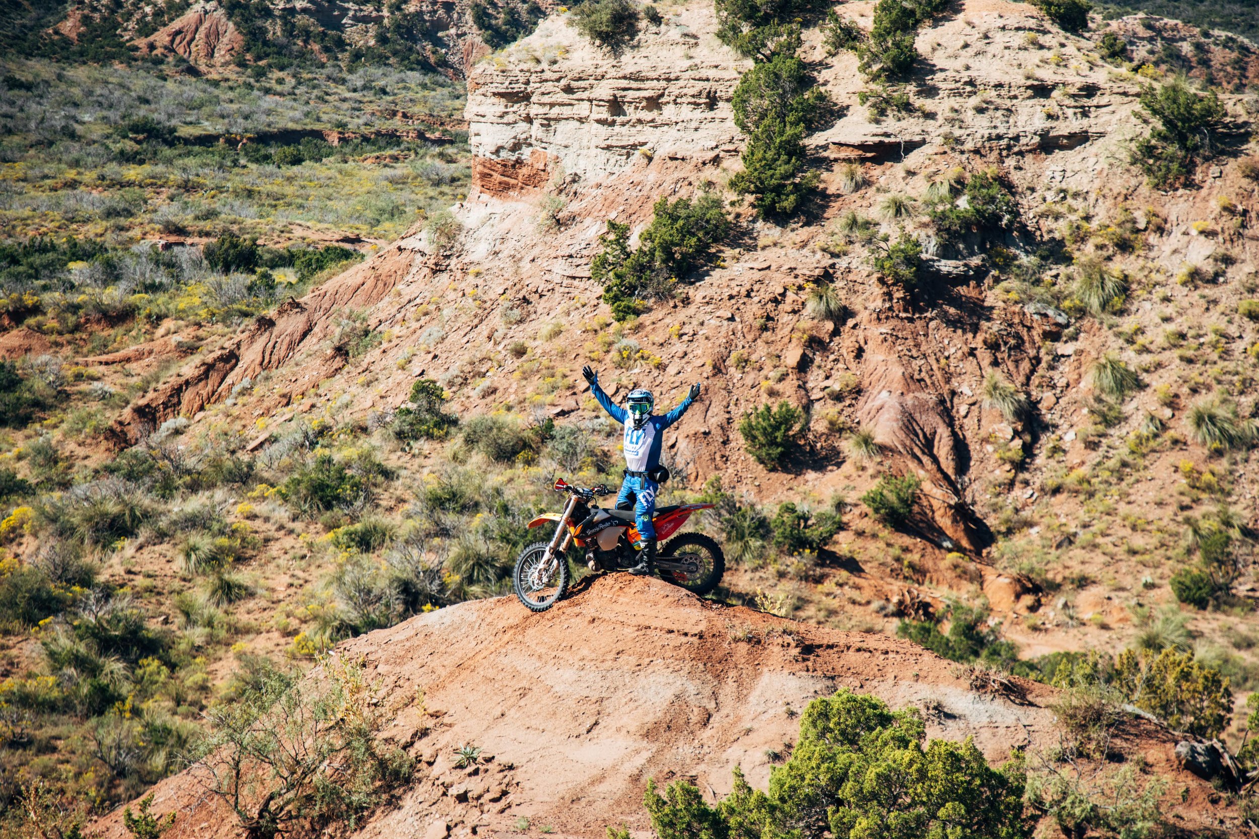 A person in motocross gear standing on a dirt trail in a desert landscape, with their arms raised, near a black and orange dirt bike, with red rock formations and sparse vegetation in the background.