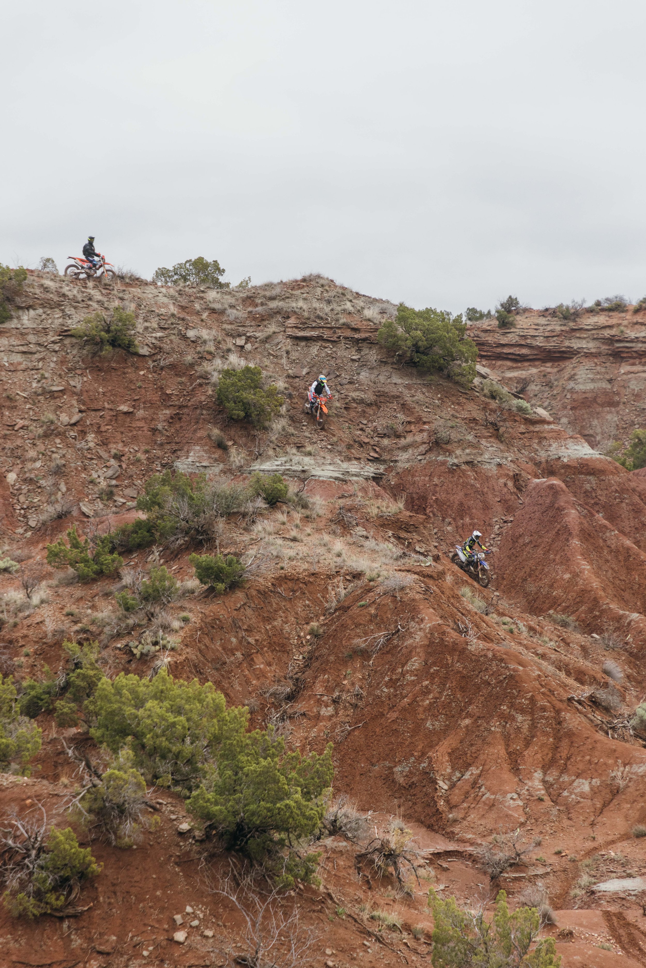 Three motorcyclists are riding down a steep, rocky dirt trail surrounded by sparse desert vegetation and small trees, with a cloudy sky overhead.