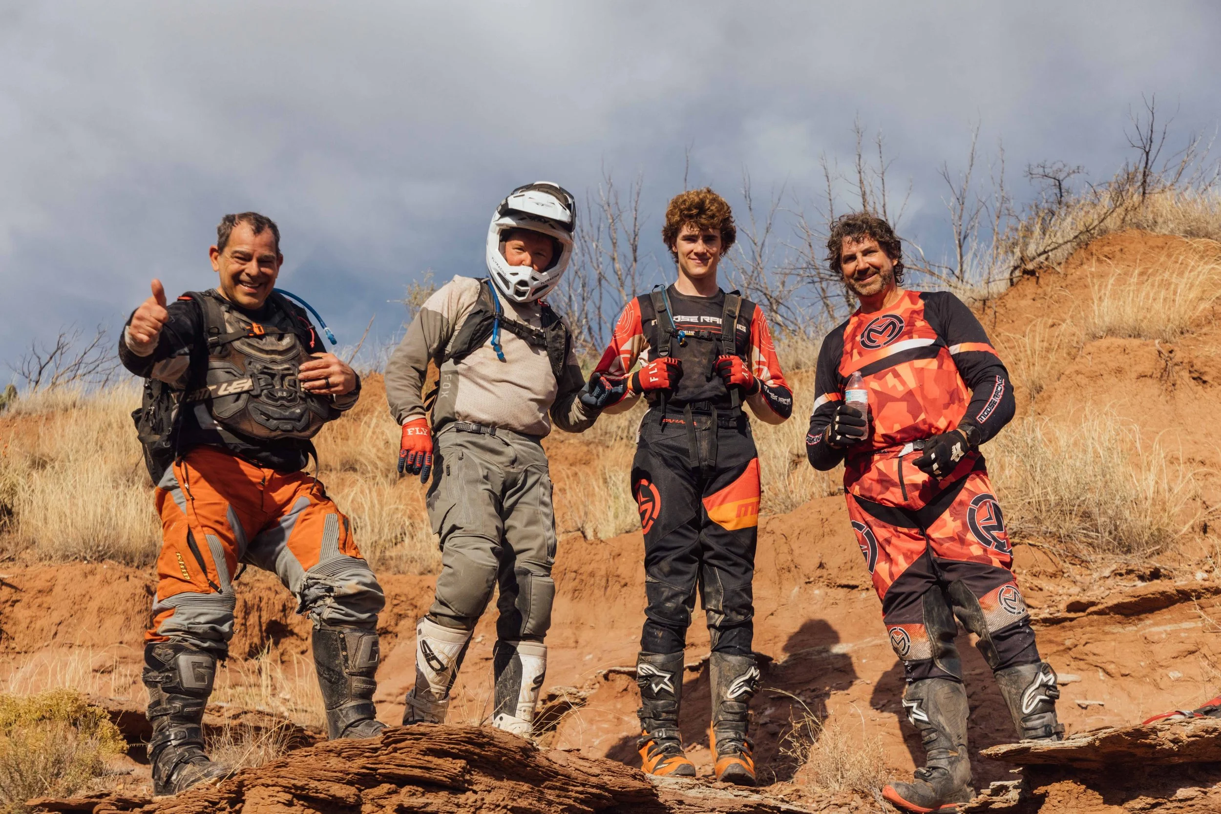 Four men standing outdoors on rocky terrain, dressed in motocross gear, smiling and posing for a photo with a cloudy sky in the background.