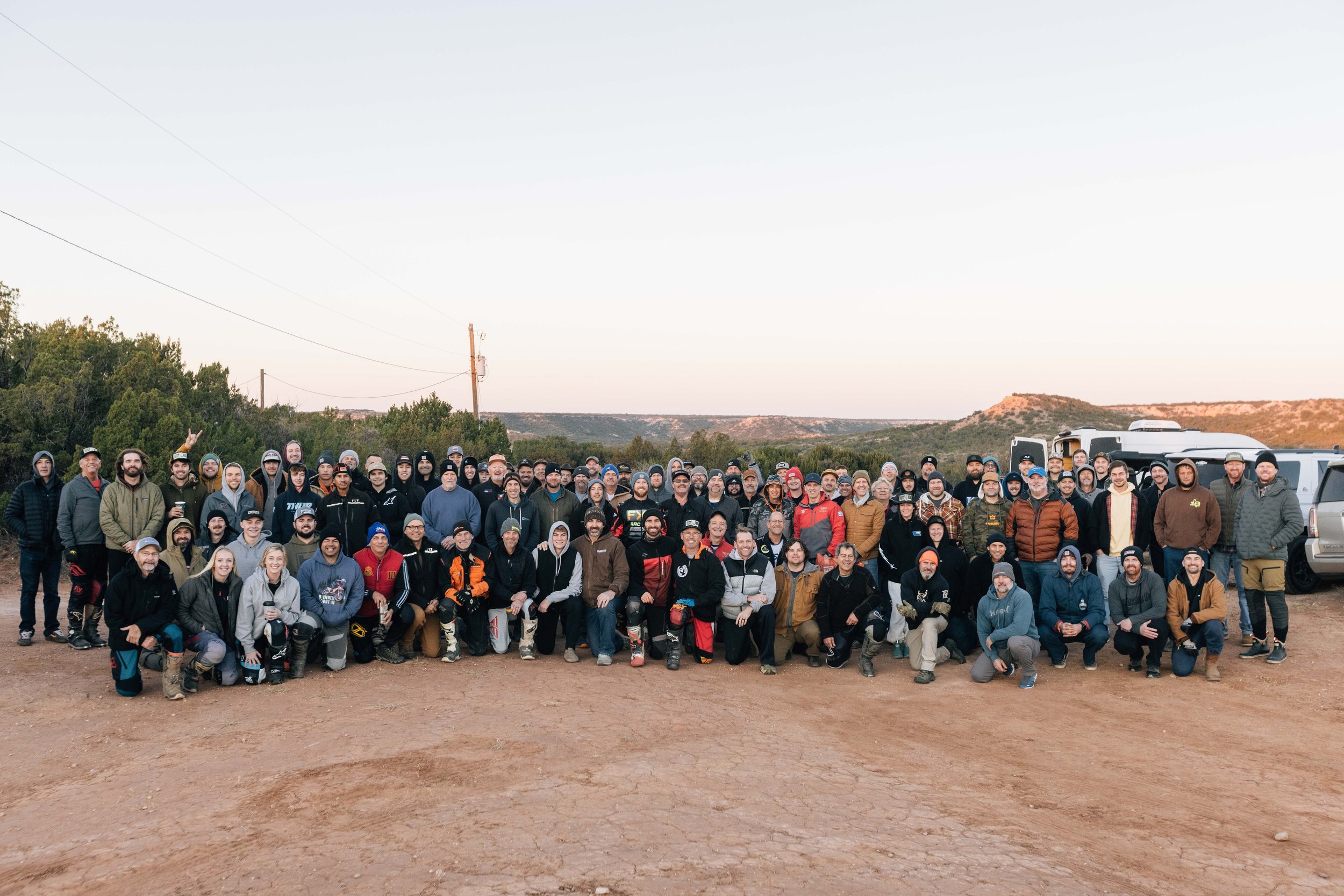 A large group of people gathered outdoors, posing for a group photo, with cars and natural landscape in the background.