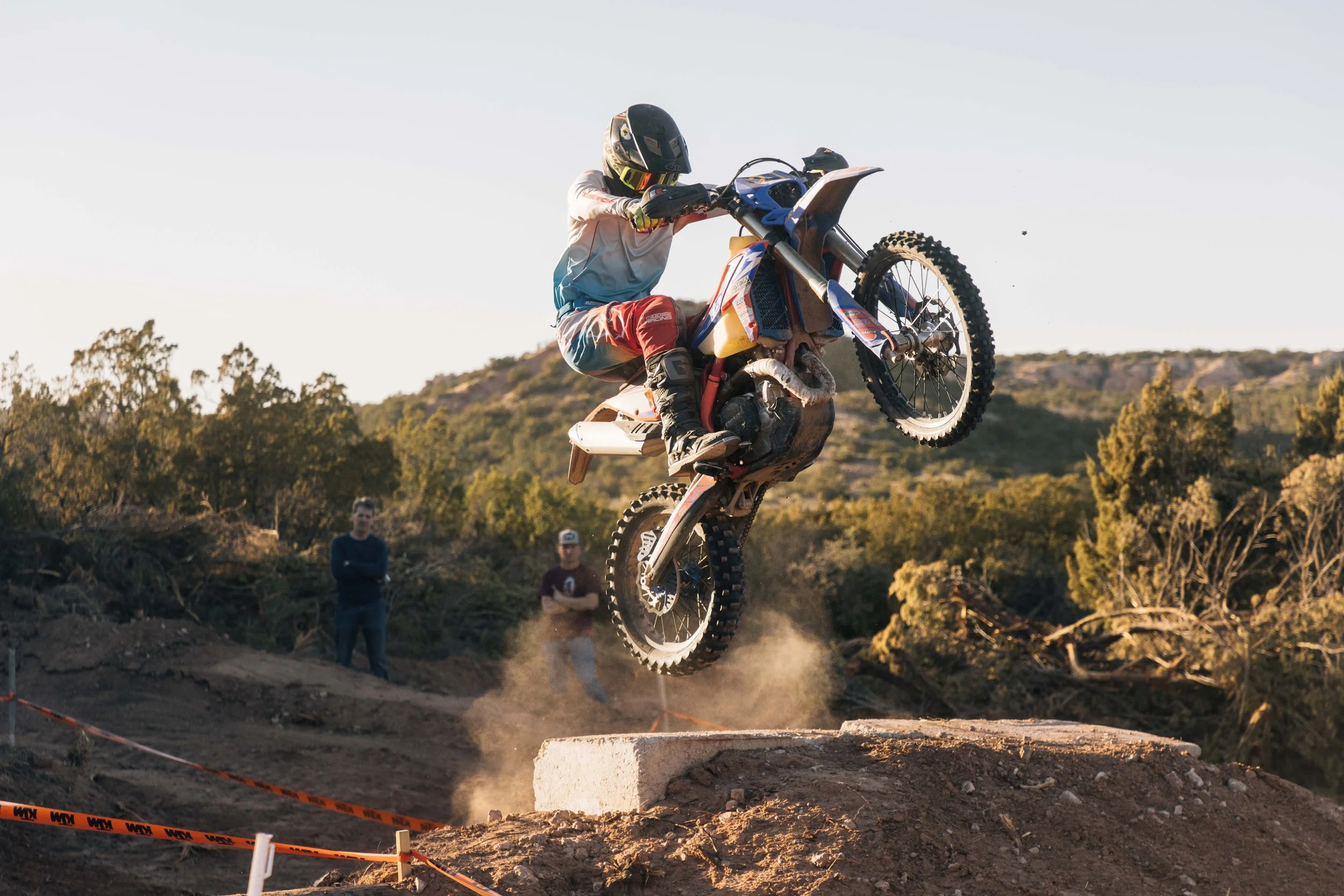 A motocross rider in a helmet and protective gear performs a jump on a dirt track, with spectators watching in the background and trees in the distance.