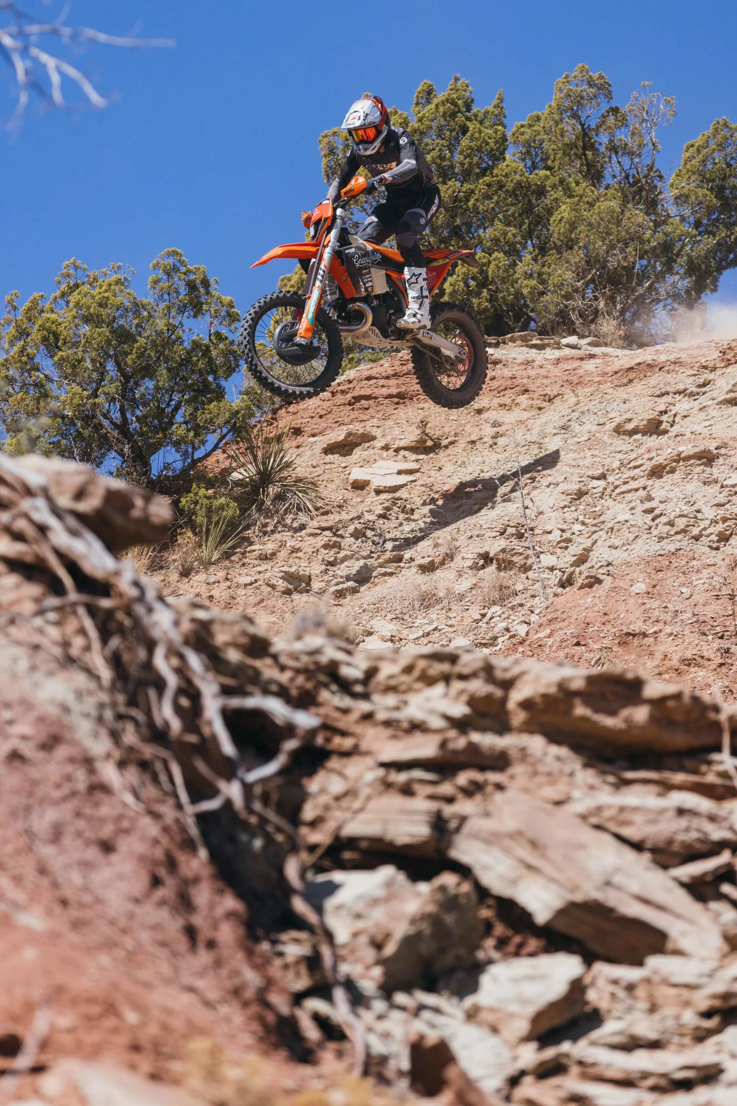 A person riding an orange motocross dirt bike on rocky terrain, mid-air, with trees and a clear blue sky in the background.