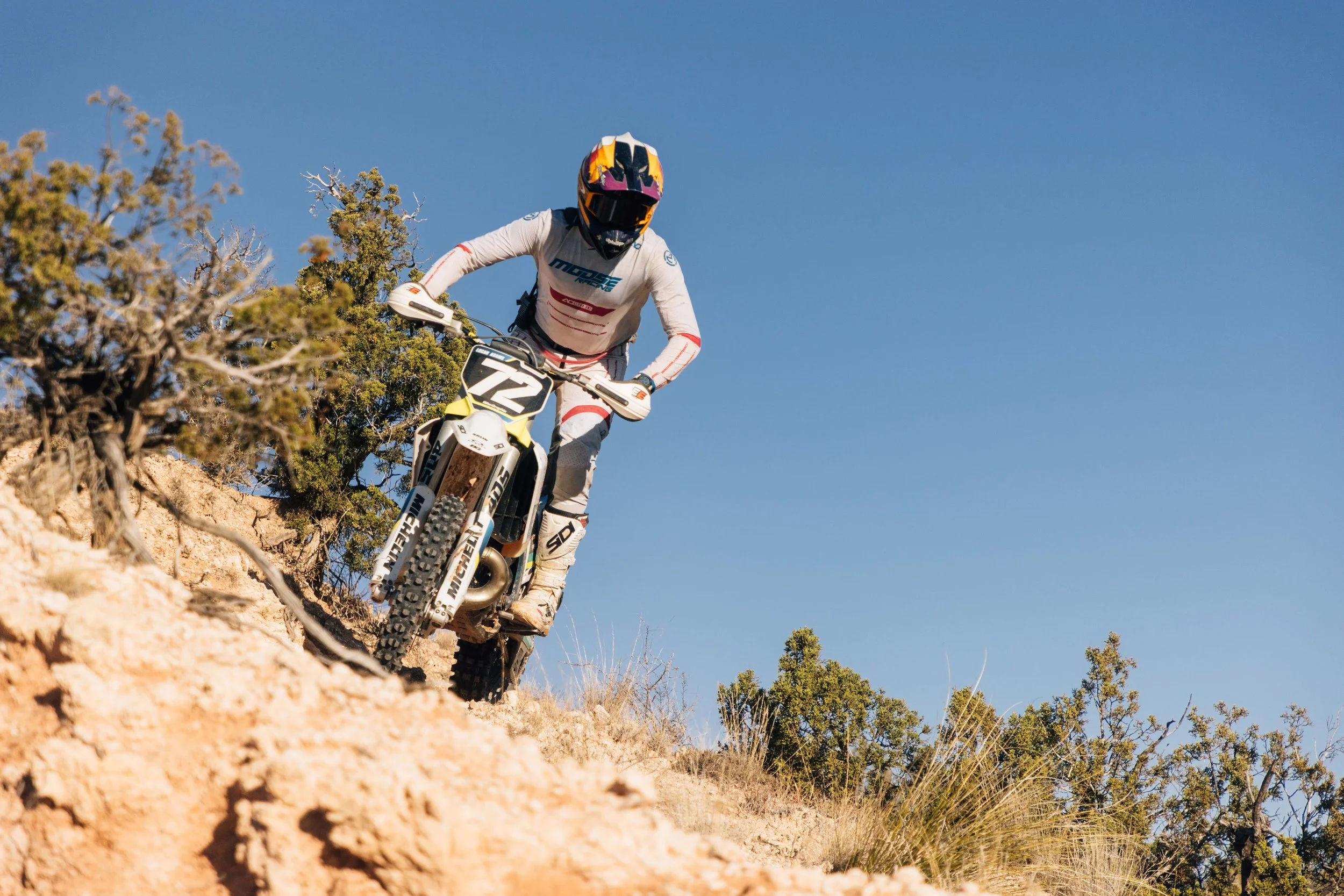 A person riding a mountain bike downhill on a dirt trail in a desert landscape with shrubs and small trees, under a clear blue sky.