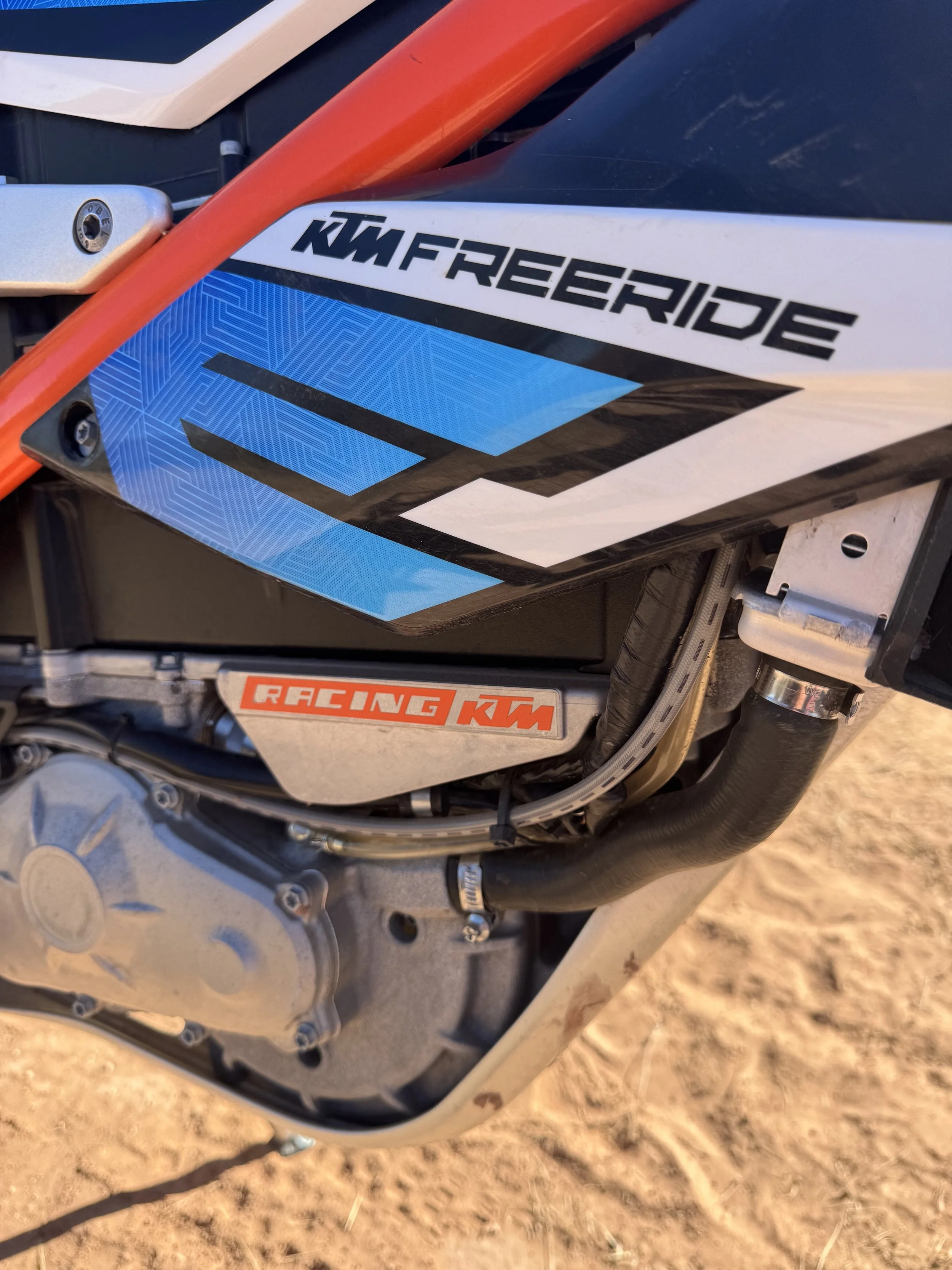 Close-up of a KTM Freeride motorcycle with a blue and white decal, showing parts of the engine, frame, and suspension, on a dirt surface.