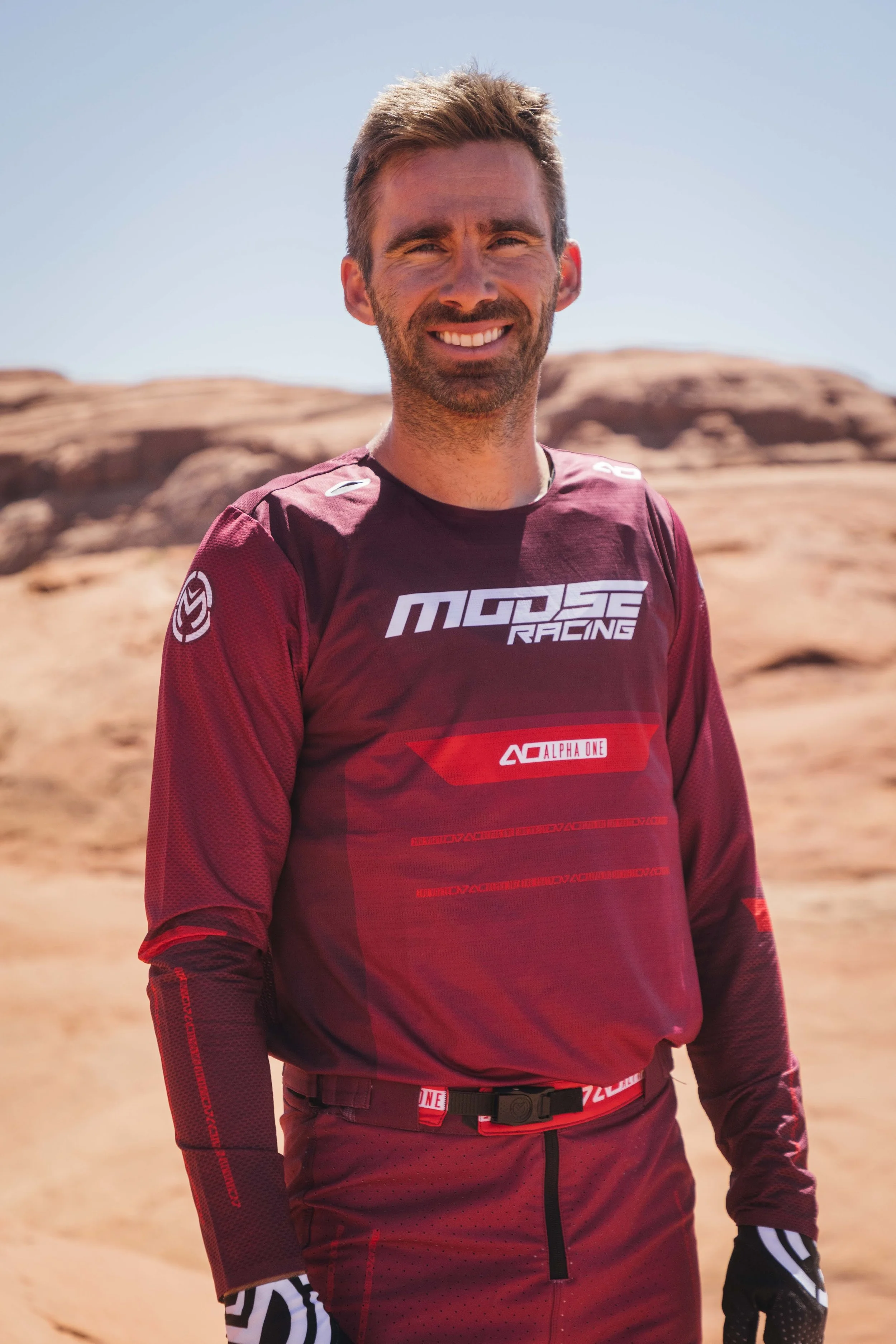 A man wearing a maroon sports jersey with 'Moose Racing' logo stands outdoors in a desert landscape with rocks and sand, smiling at the camera.