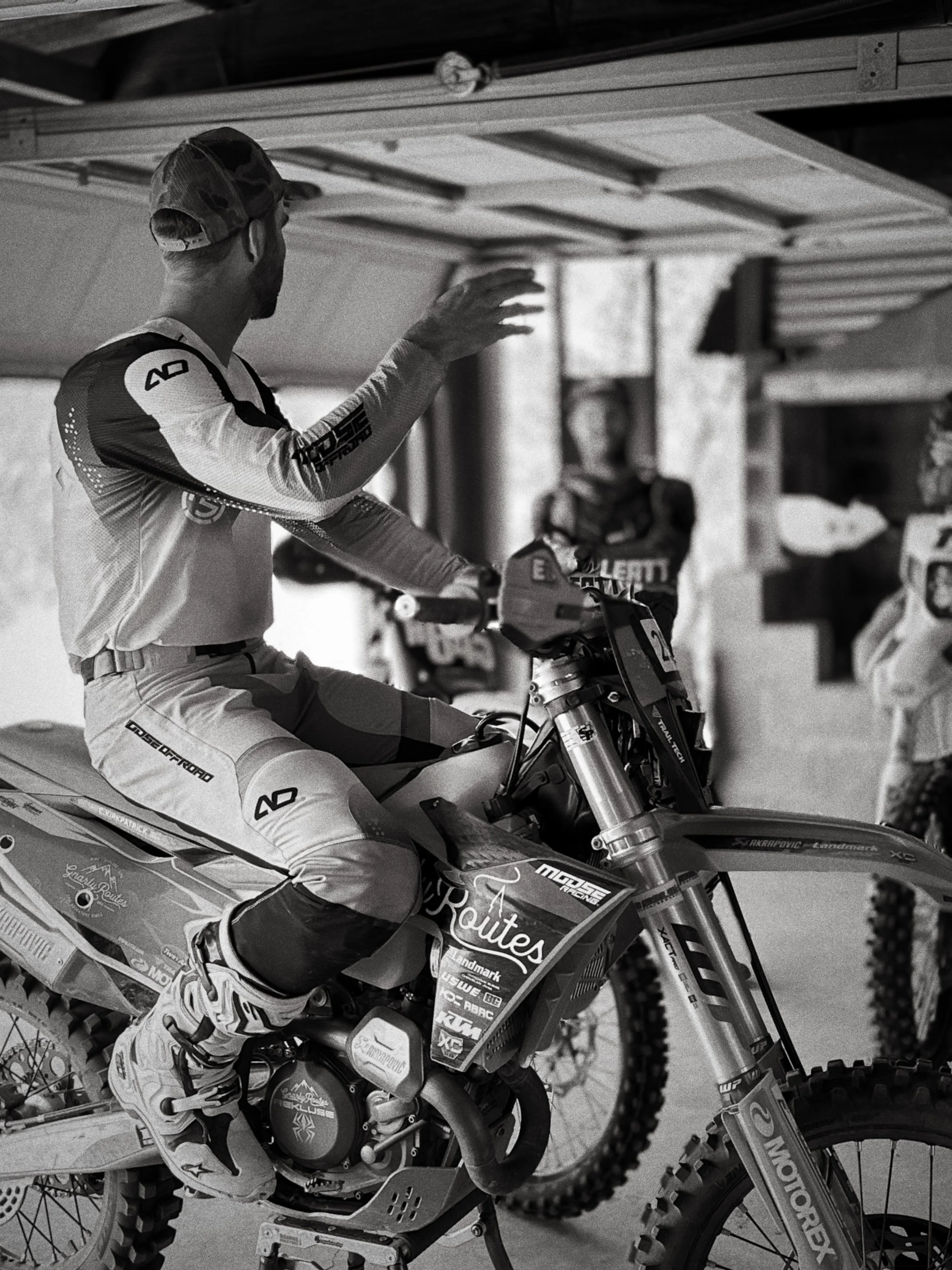 A person dressed in motocross gear sitting on a dirt bike, with other riders and equipment in the background, in black and white.