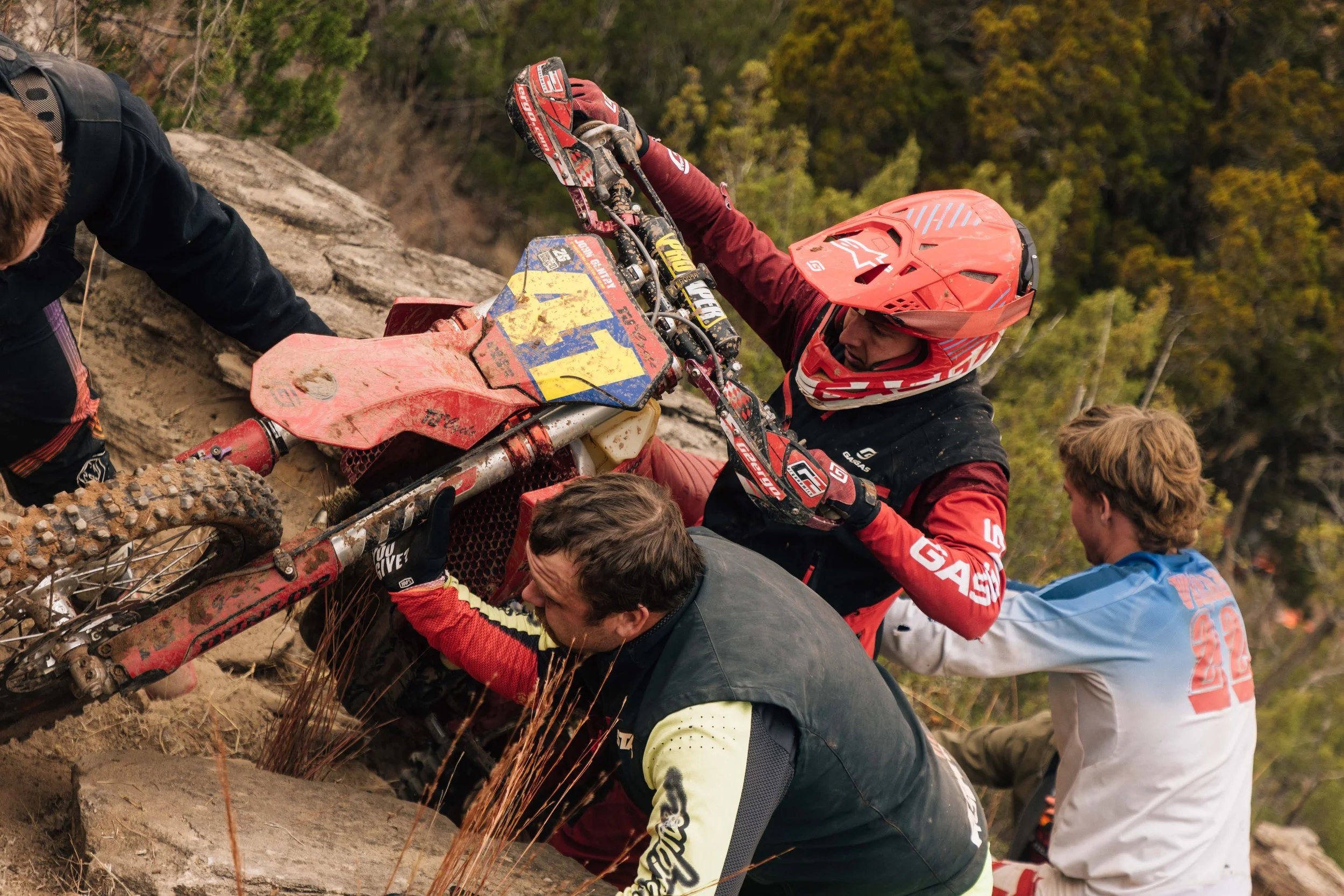 Motocross riders assisting a fallen rider on a dirt trail in a forested area during a race.