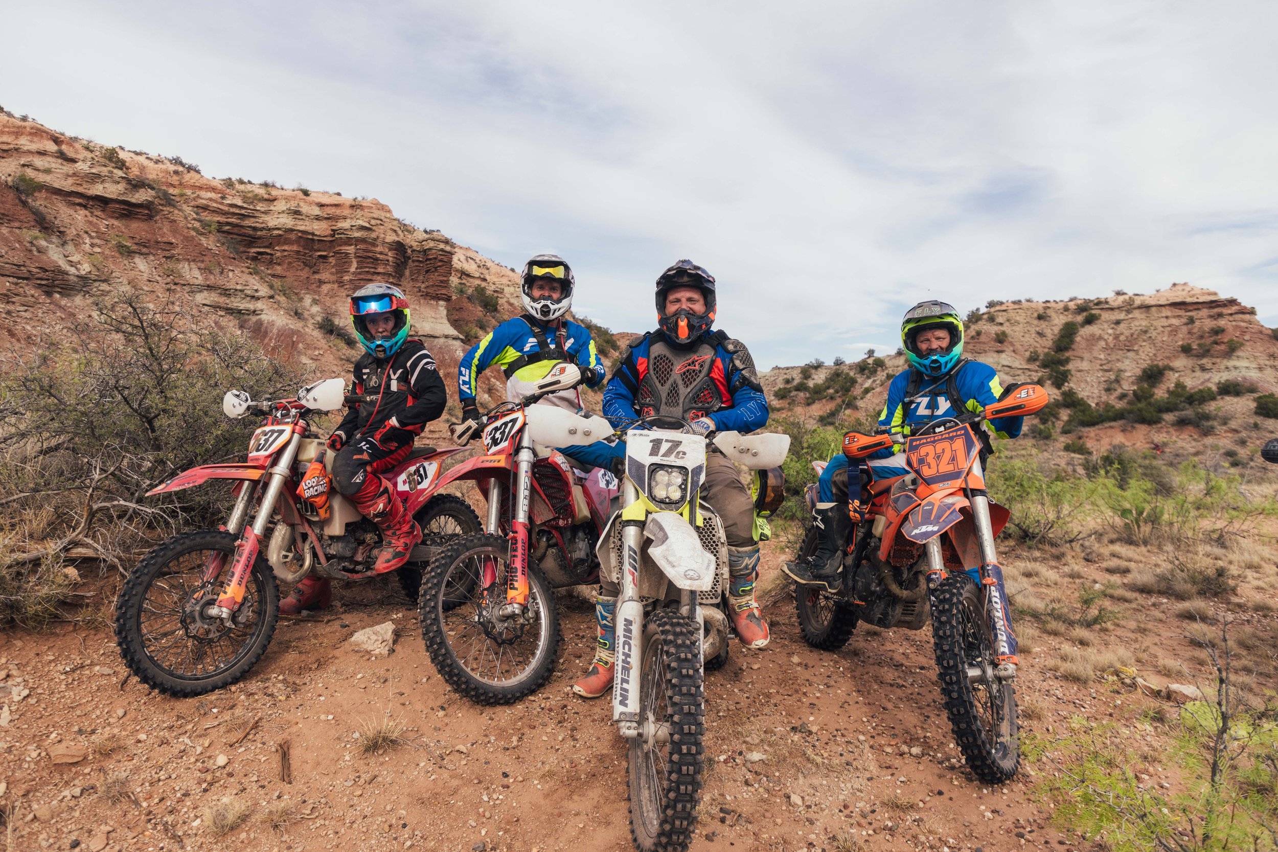 Four motocross riders in full gear posing with their dirt bikes on a dirt trail with a rocky, desert hillside in the background.