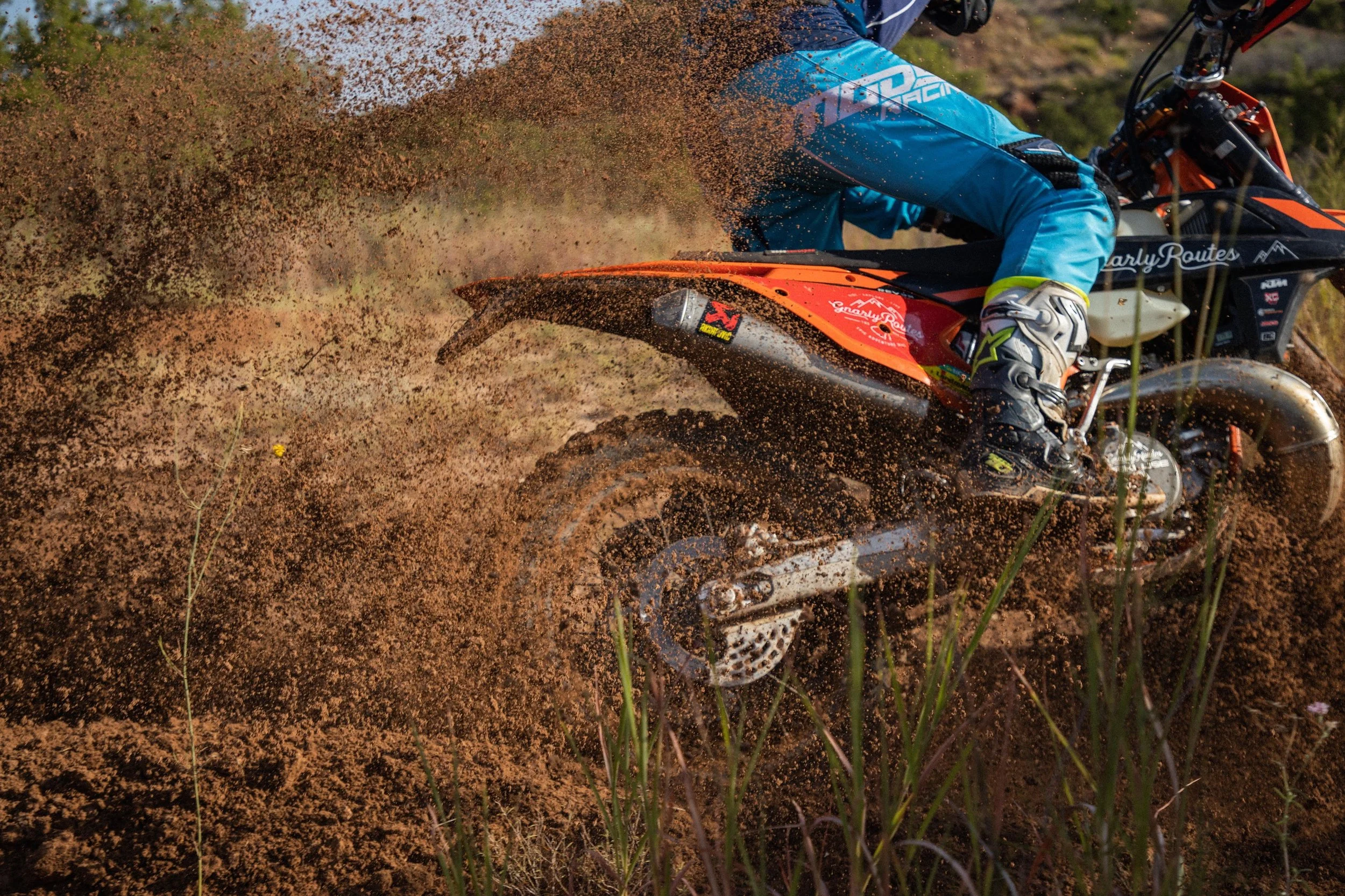 A dirt bike rider wearing a blue jacket and helmet, leaning into a turn on a dirt trail, kicking up a cloud of reddish dirt and small rocks.