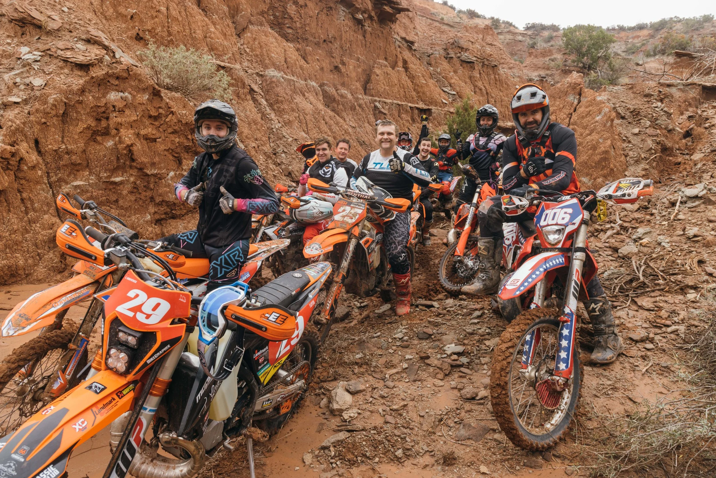 A group of motocross riders in gear and helmets, smiling and giving thumbs up, sitting and standing around their dirt bikes on a rocky dirt trail with reddish cliffs and sparse vegetation in the background.