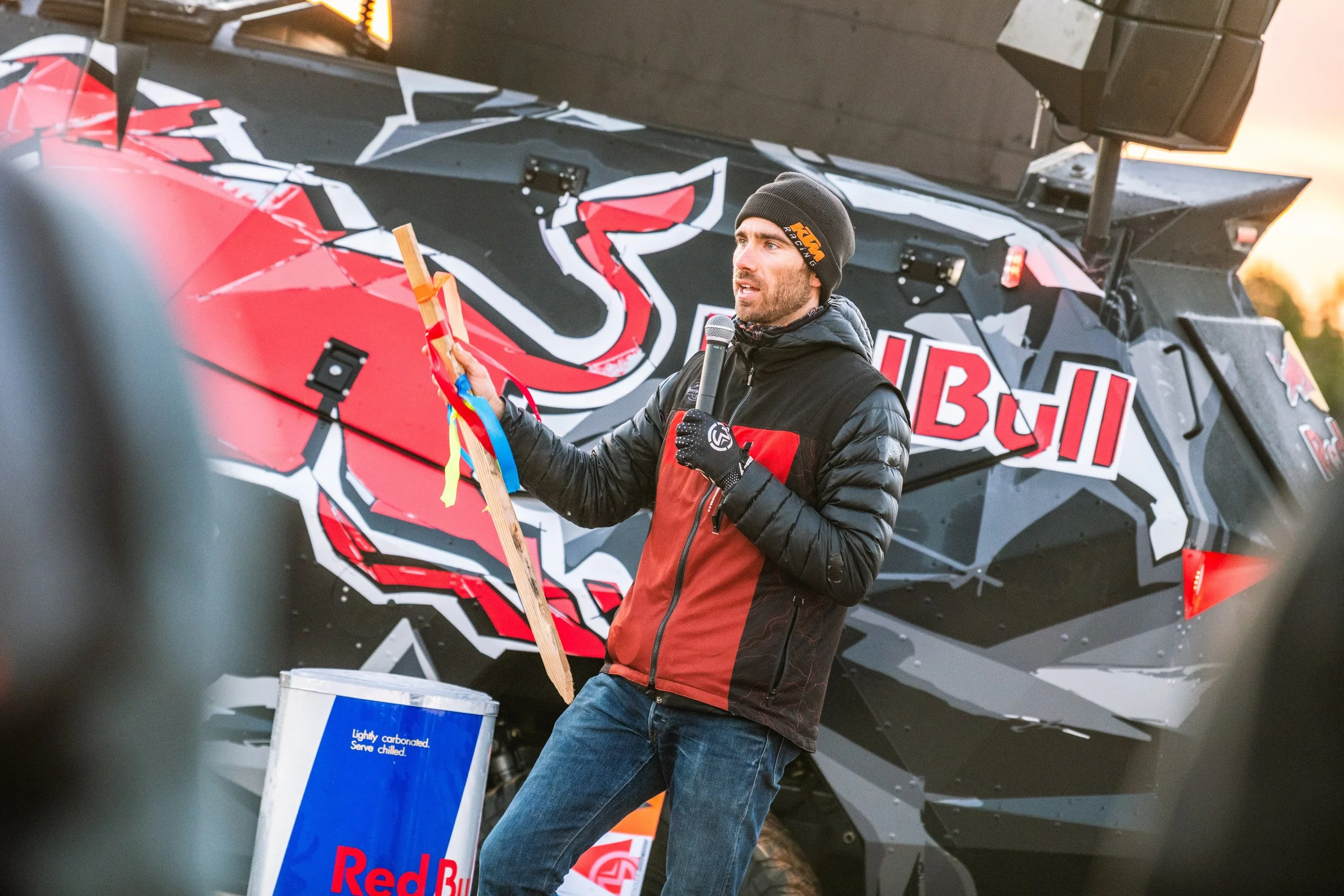 A man wearing a black and red jacket, beanie, and gloves speaking into a microphone, standing in front of a large Red Bull Racing vehicle with bold black, red, white, and grey graphics, and holding a wooden stick with colorful ribbons attached.