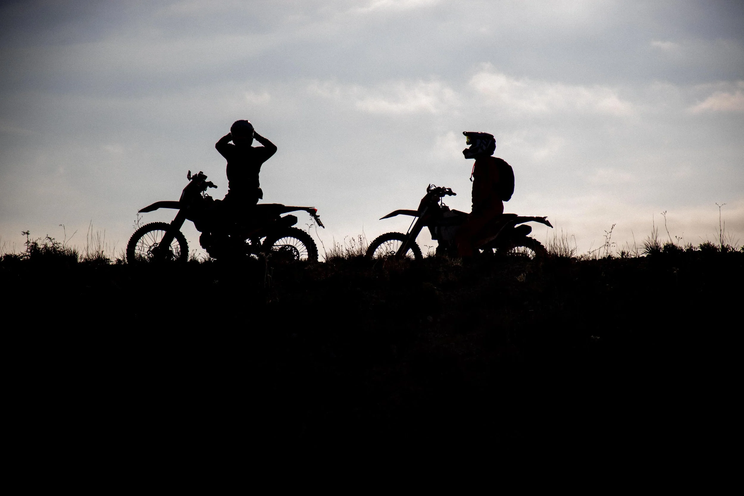 Two people with backpacks and helmets sitting on dirt bikes on a hilltop during sunset.