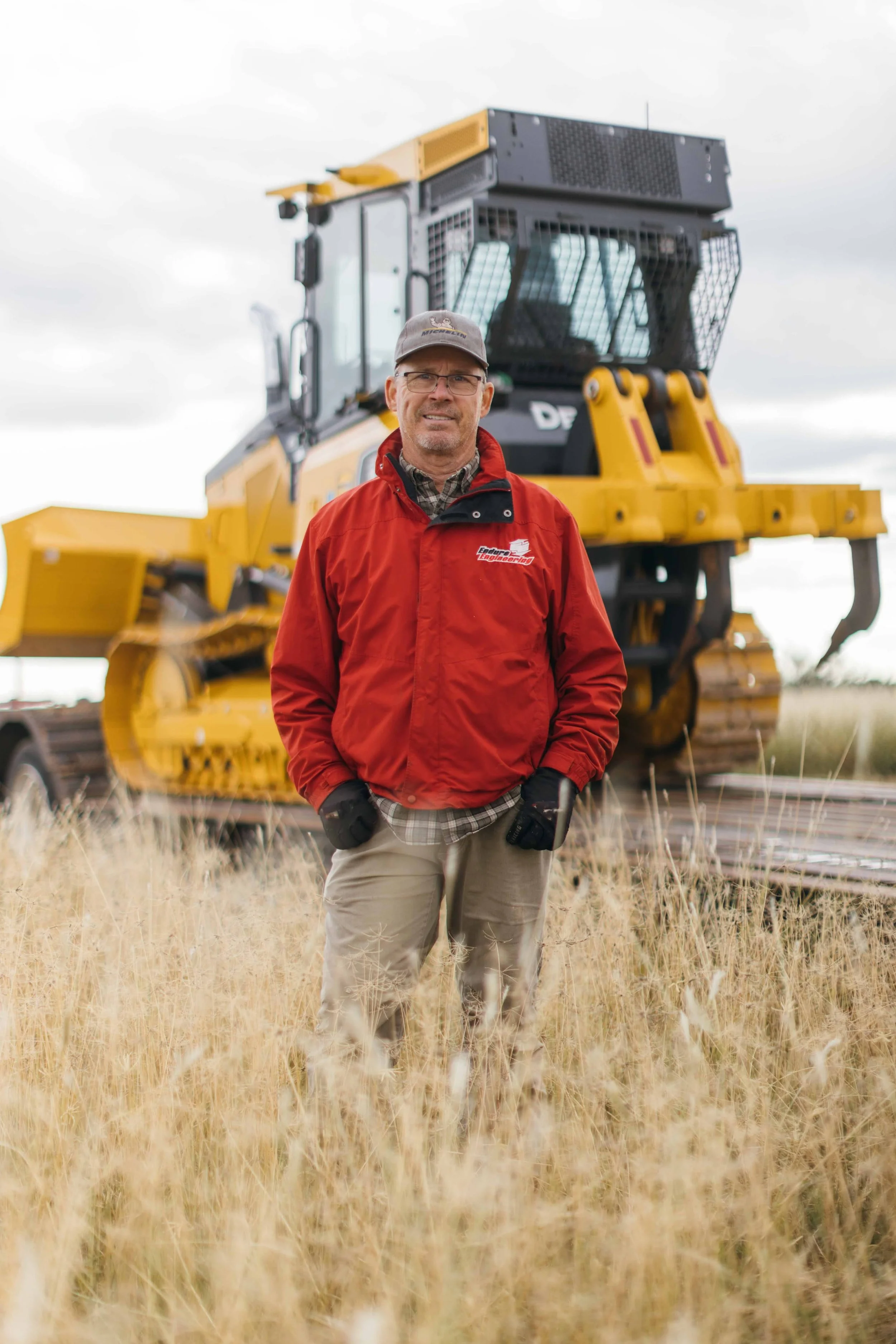 A man wearing glasses, a red jacket, and a gray cap standing in a field of tall, dry grass with a large yellow construction machine in the background under cloudy skies.