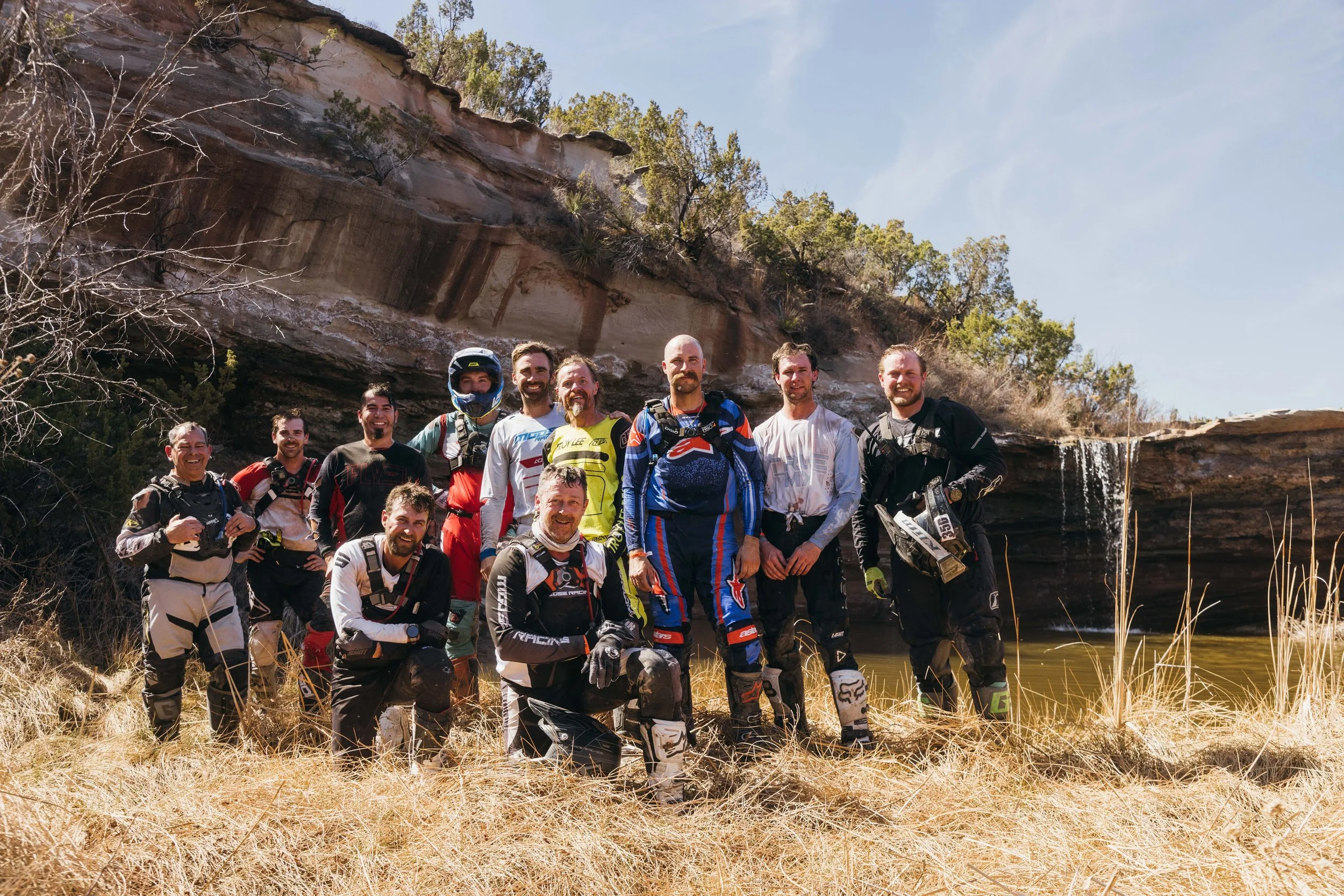 A group of male motorcycle dirt bikers in gear standing together outdoors in front of a rocky cliff and a shallow body of water with dry grass in the foreground.