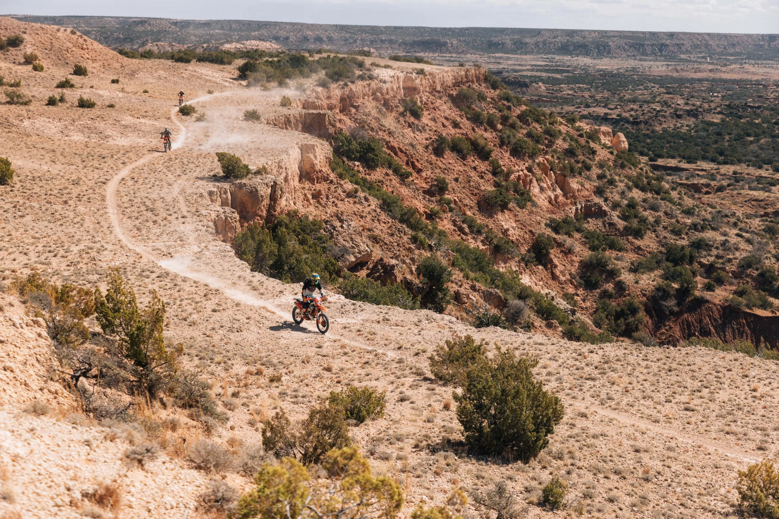Multiple off-road motorcyclists riding on a dusty trail through a desert landscape with sparse vegetation and rugged terrain.