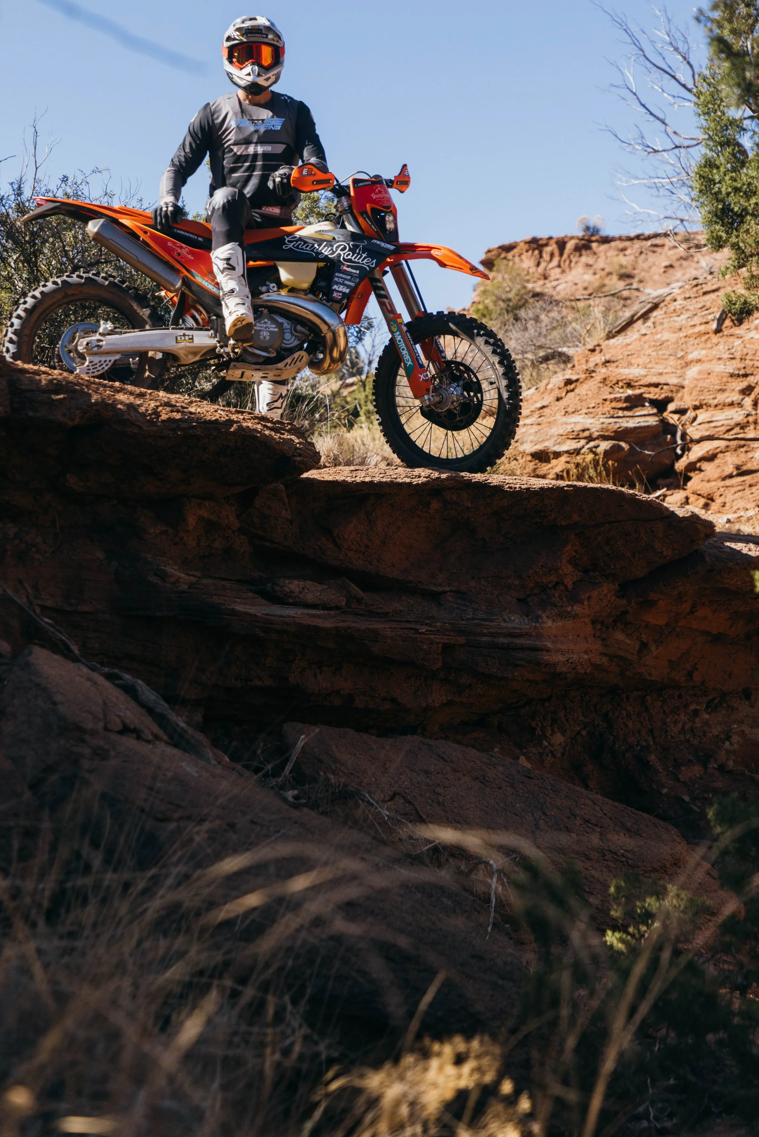 A person wearing motocross gear, including helmet and goggles, riding an orange dirt bike on rugged rocky terrain in a desert landscape with sparse vegetation and red rock formations.